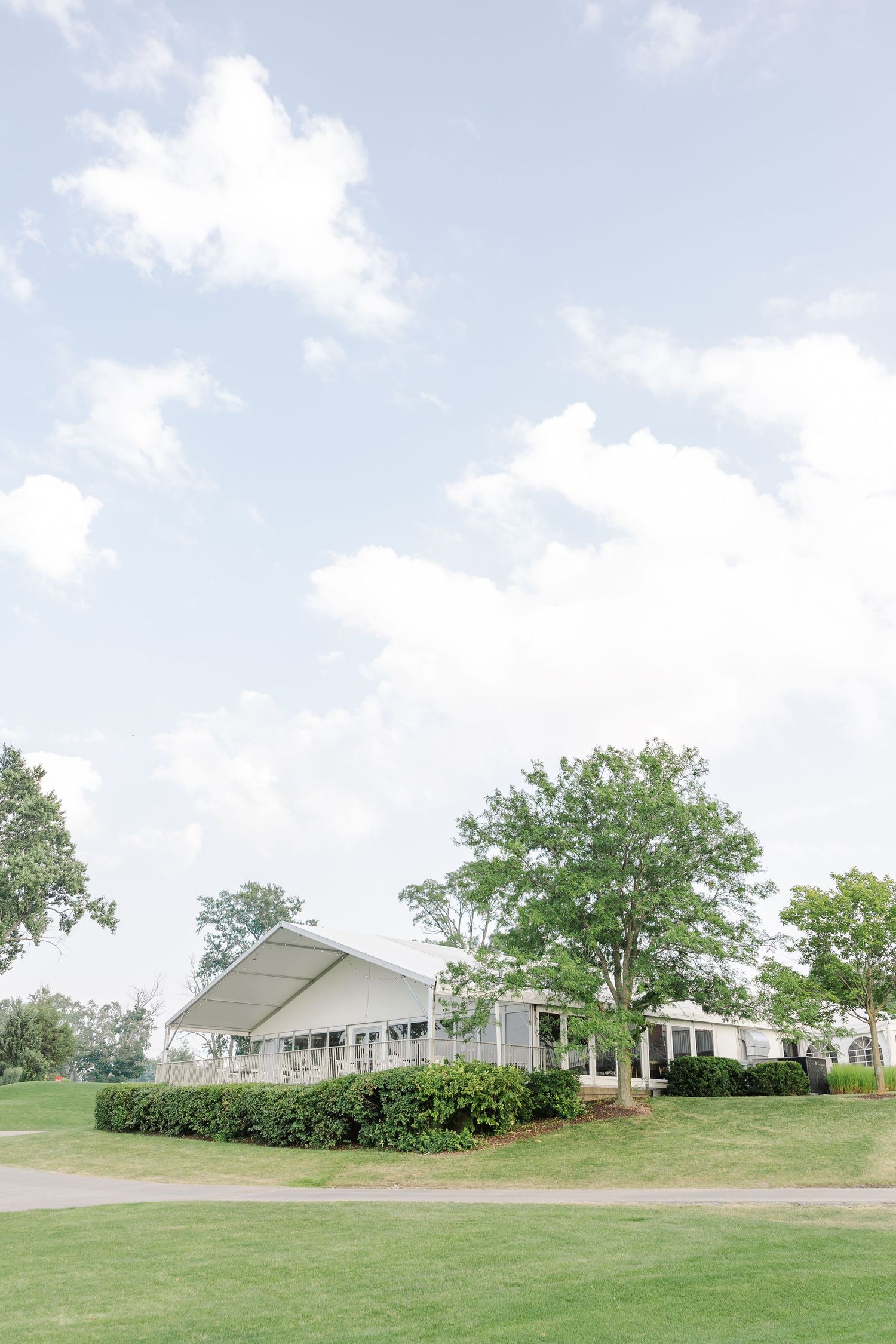 White wedding venue building surrounded by lush green trees and manicured lawn under cloudy sky.