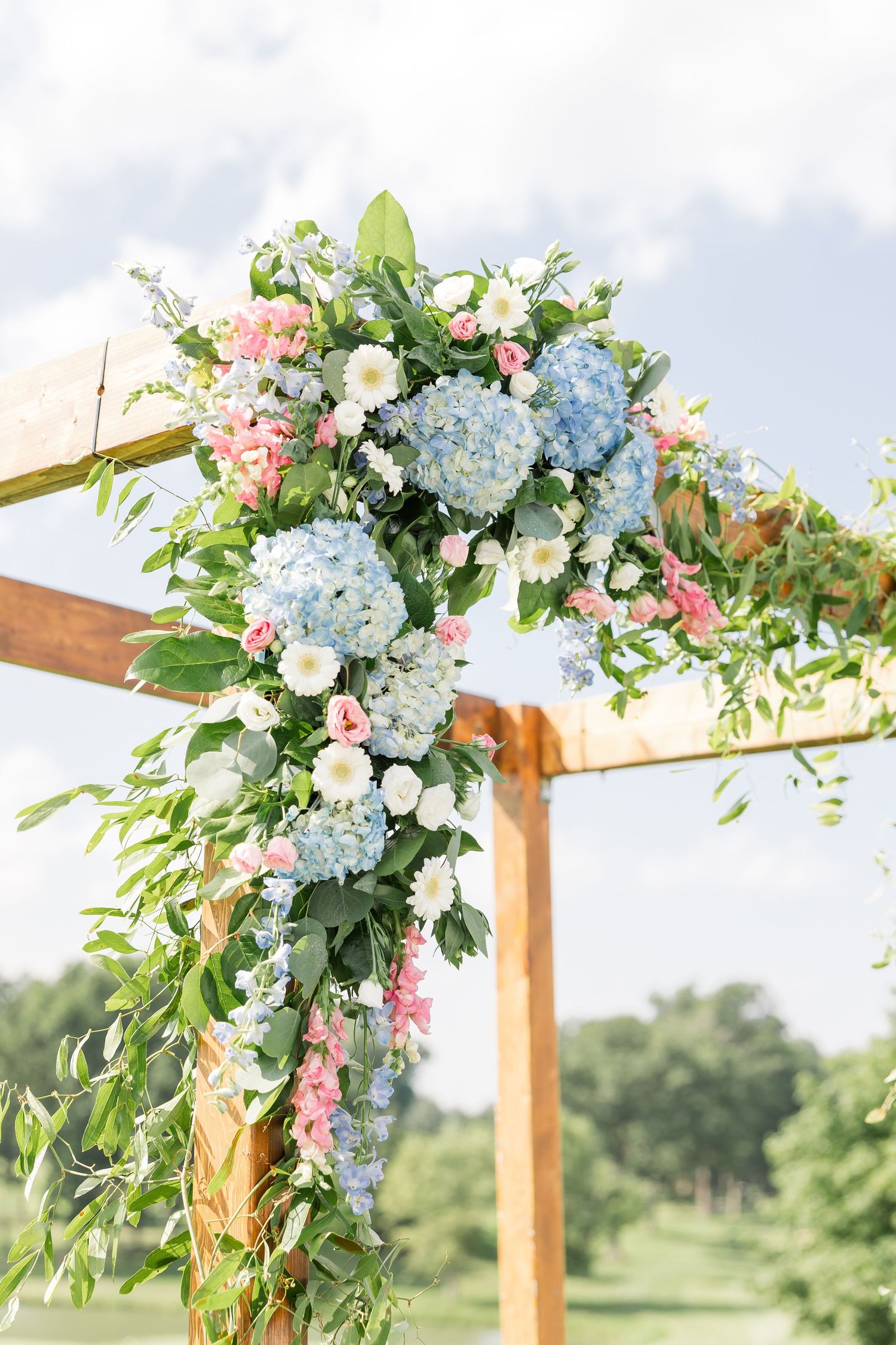 Rustic wooden wedding arch adorned with blue hydrangeas, pink and white flowers, and lush greenery outdoors.