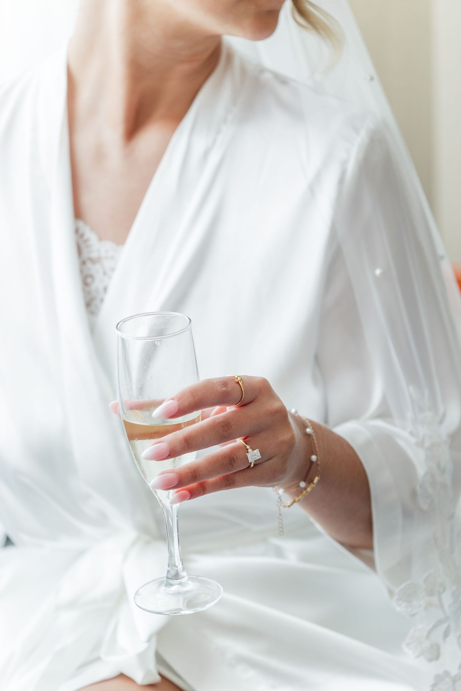 Bride in white satin robe holding champagne flute with diamond engagement ring and gold bracelet on wedding day.