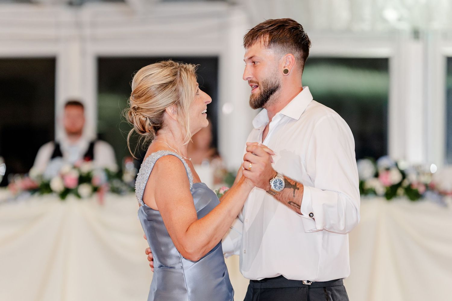 Groom dancing with mother in silver dress during wedding reception in elegant venue.
