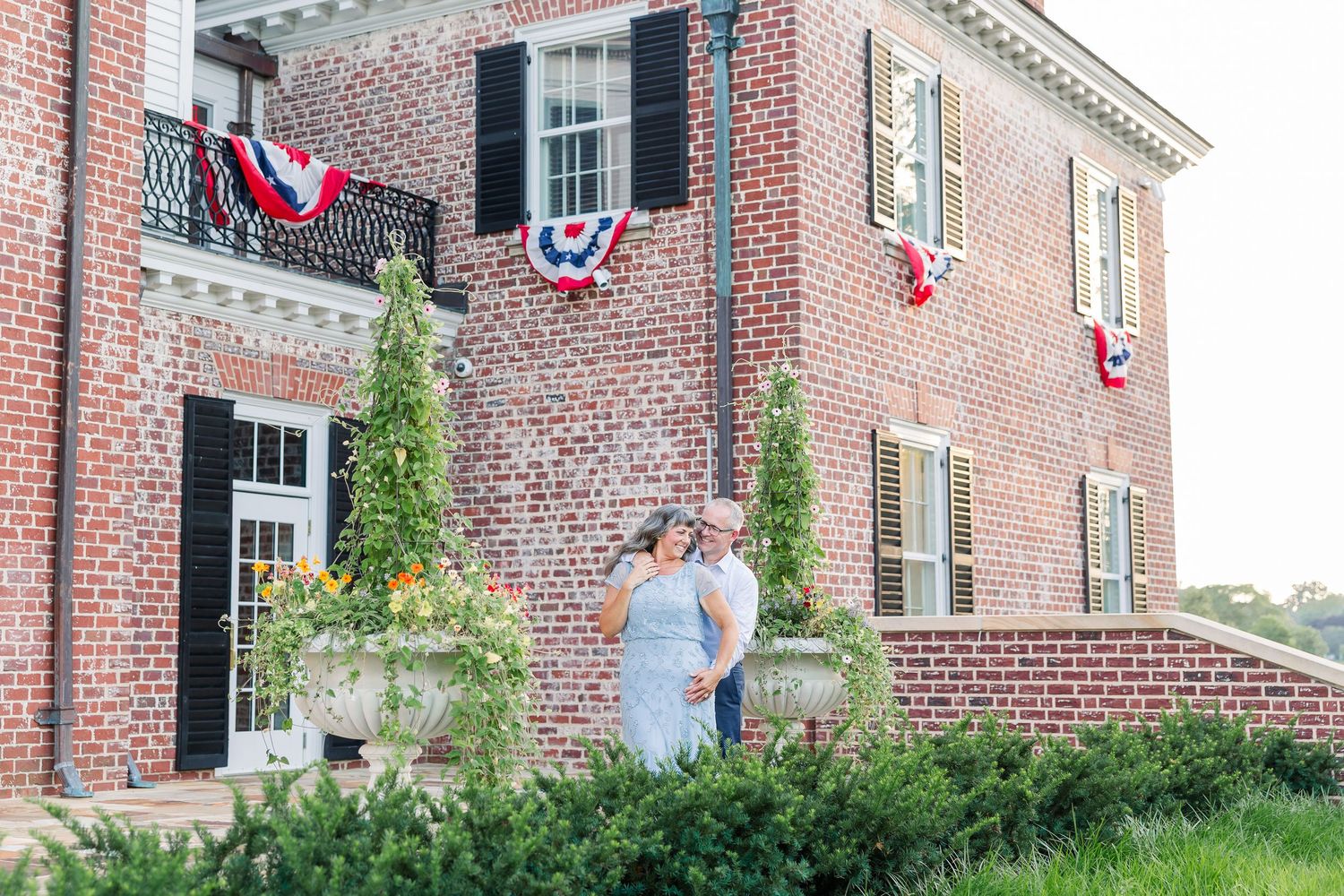 Couple stands before a brick colonial mansion adorned with patriotic red, white, and blue bunting decorations.