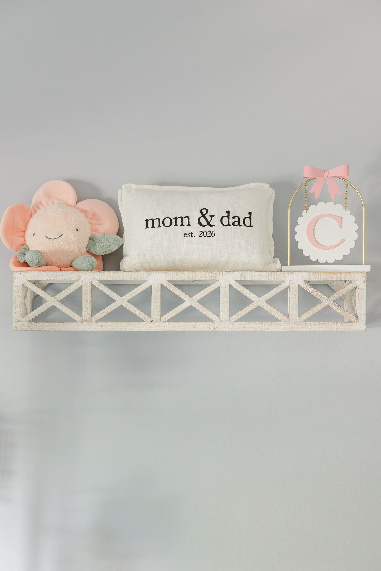 White nursery shelf with plush flower toy, mom & dad est. 2026 pillow, and pink monogram letter C decoration.