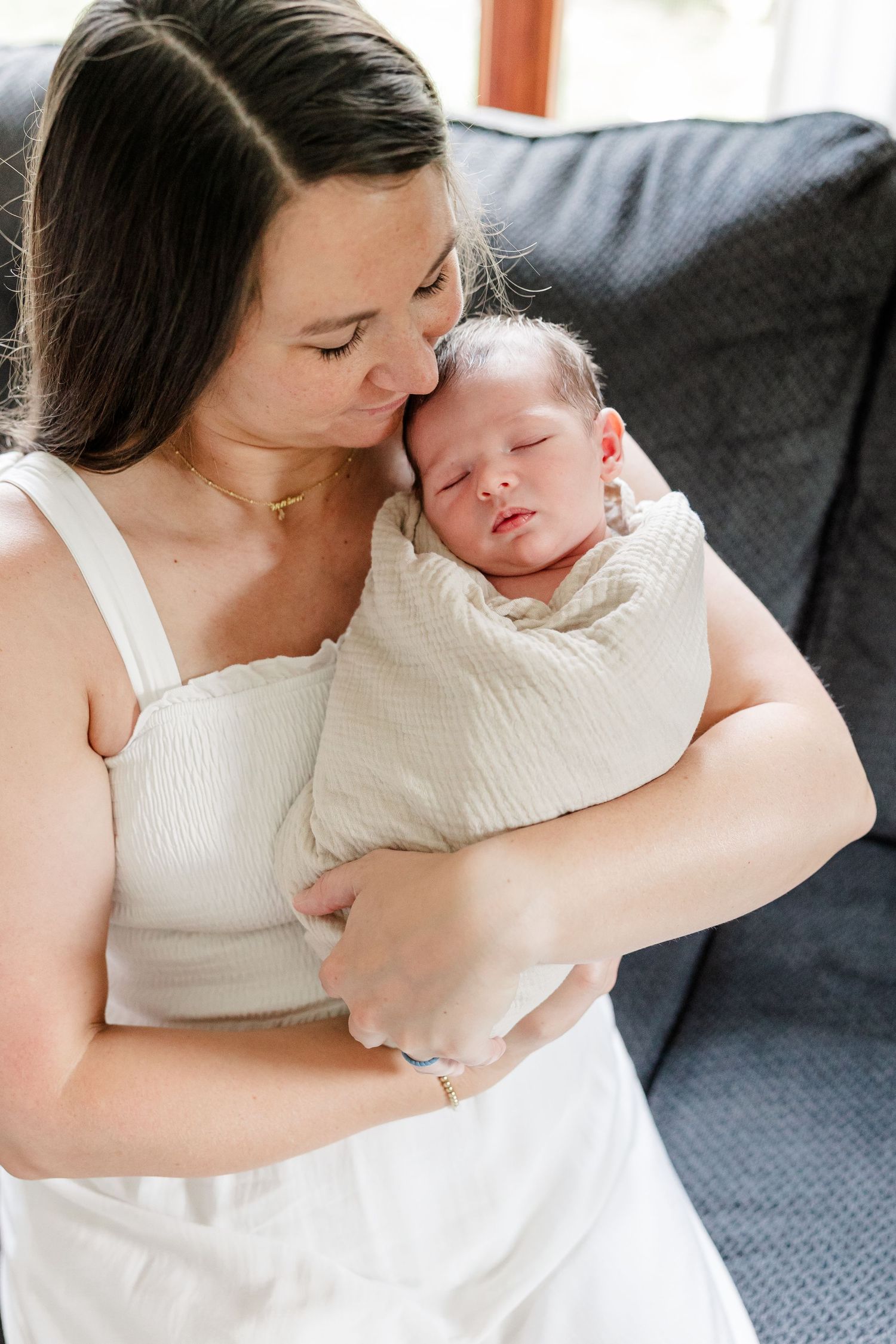 New mother in white dress tenderly holding her swaddled sleeping newborn baby close to her cheek.