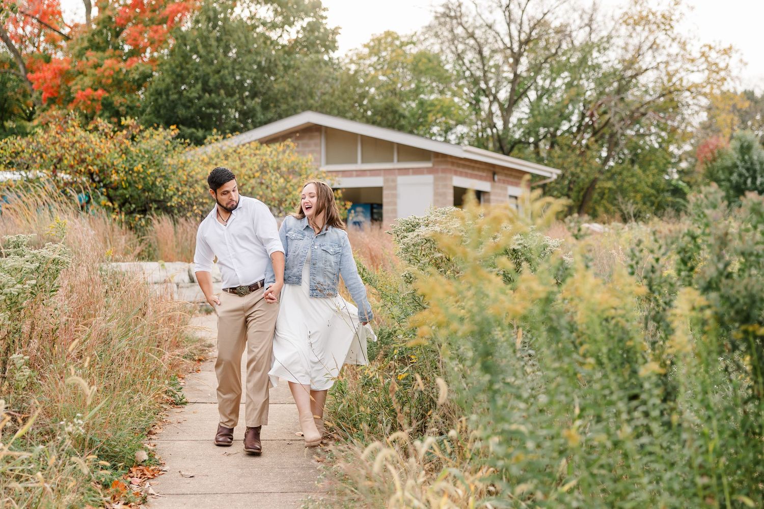 Smiling couple in casual clothes walking hand in hand along a garden path lined with tall grasses and autumn foliage near a small building.