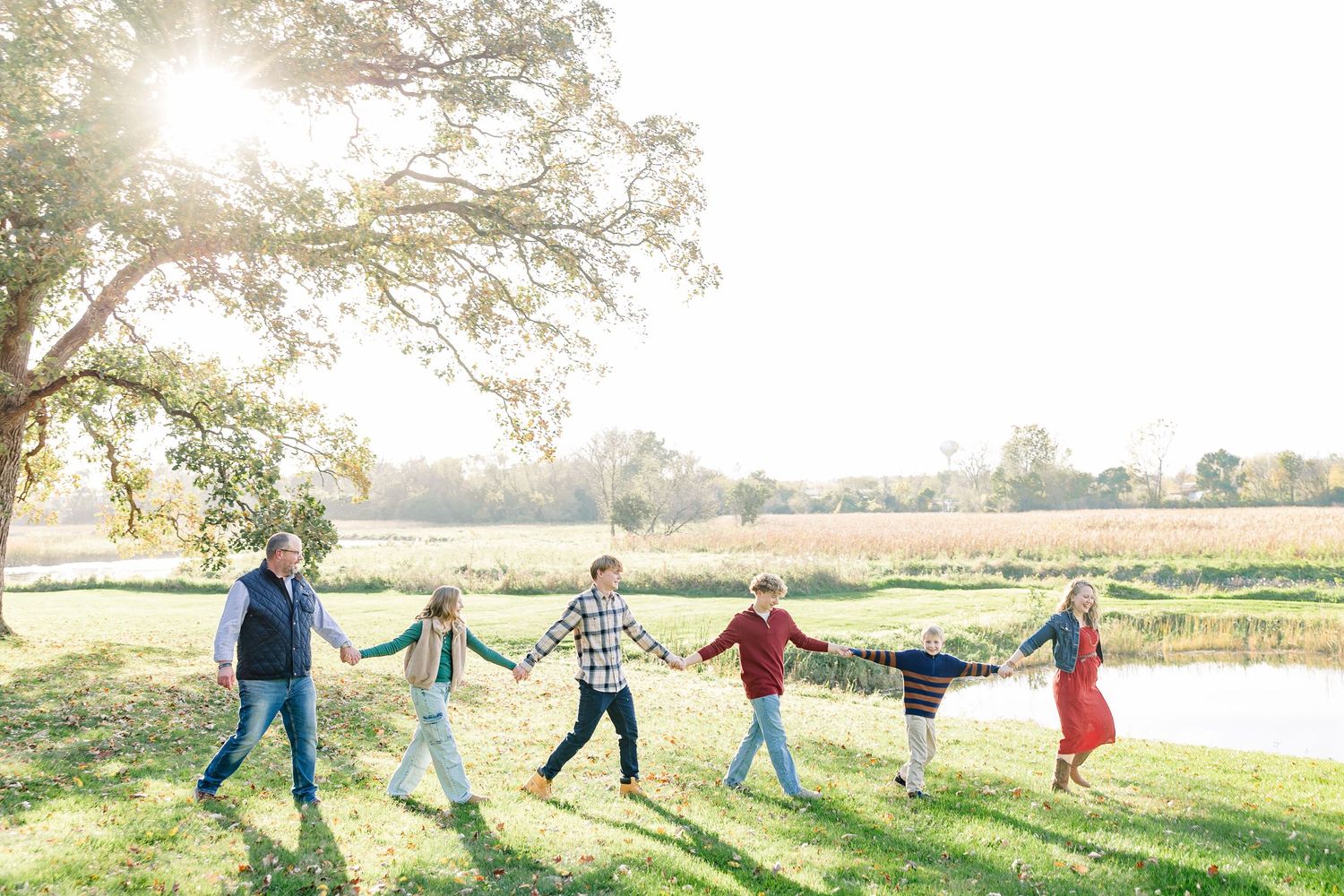 A family of six holds hands in a line while walking across a sunlit grassy field near a pond, with warm autumn light behind them.