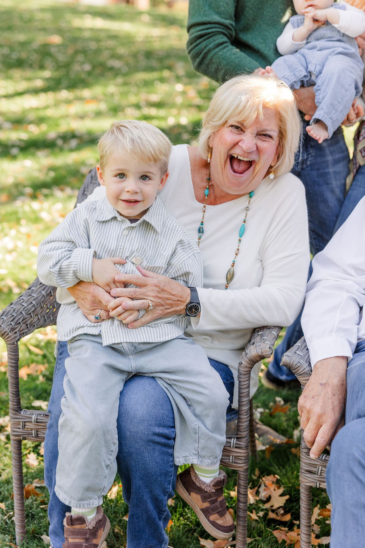 Grandmother laughing joyfully while holding young blonde boy on her lap during outdoor family photo session.