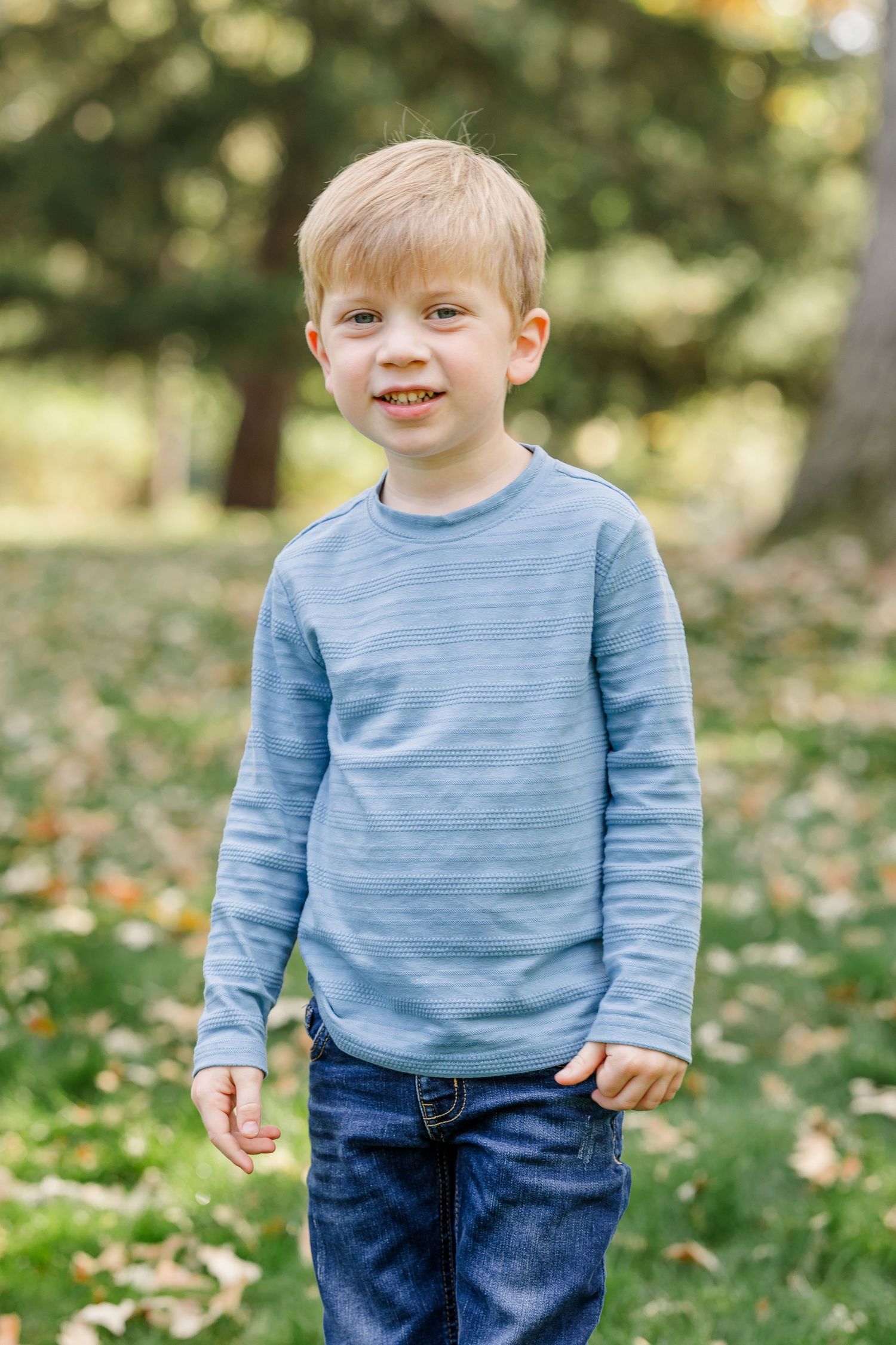 A smiling young blonde boy wearing a blue striped long-sleeve shirt and jeans standing outdoors in a park.