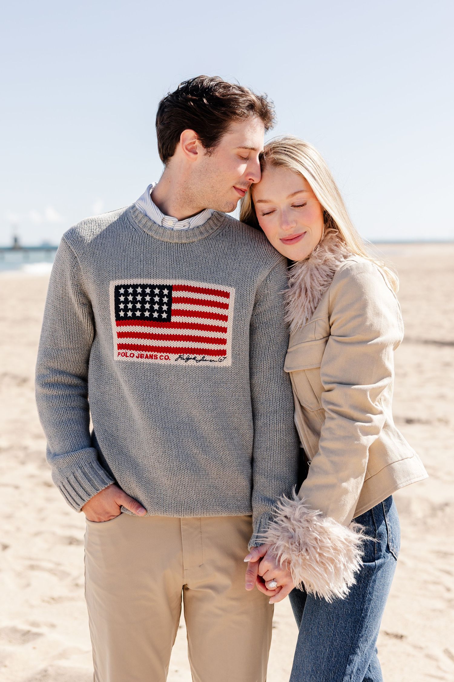 Couple embraces on beach, man wearing Polo Ralph Lauren American flag sweater.