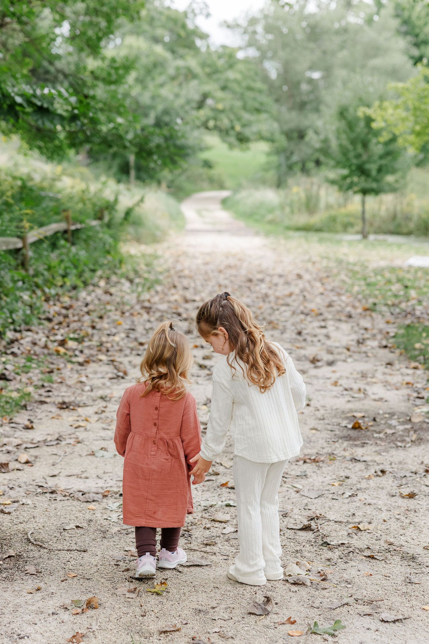 Two young sisters hold hands while walking down a scenic tree-lined dirt path in autumn.