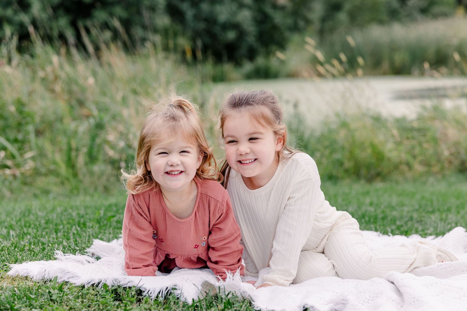 Two smiling young girls lying on a blanket in green grass near a pond on a sunny day.