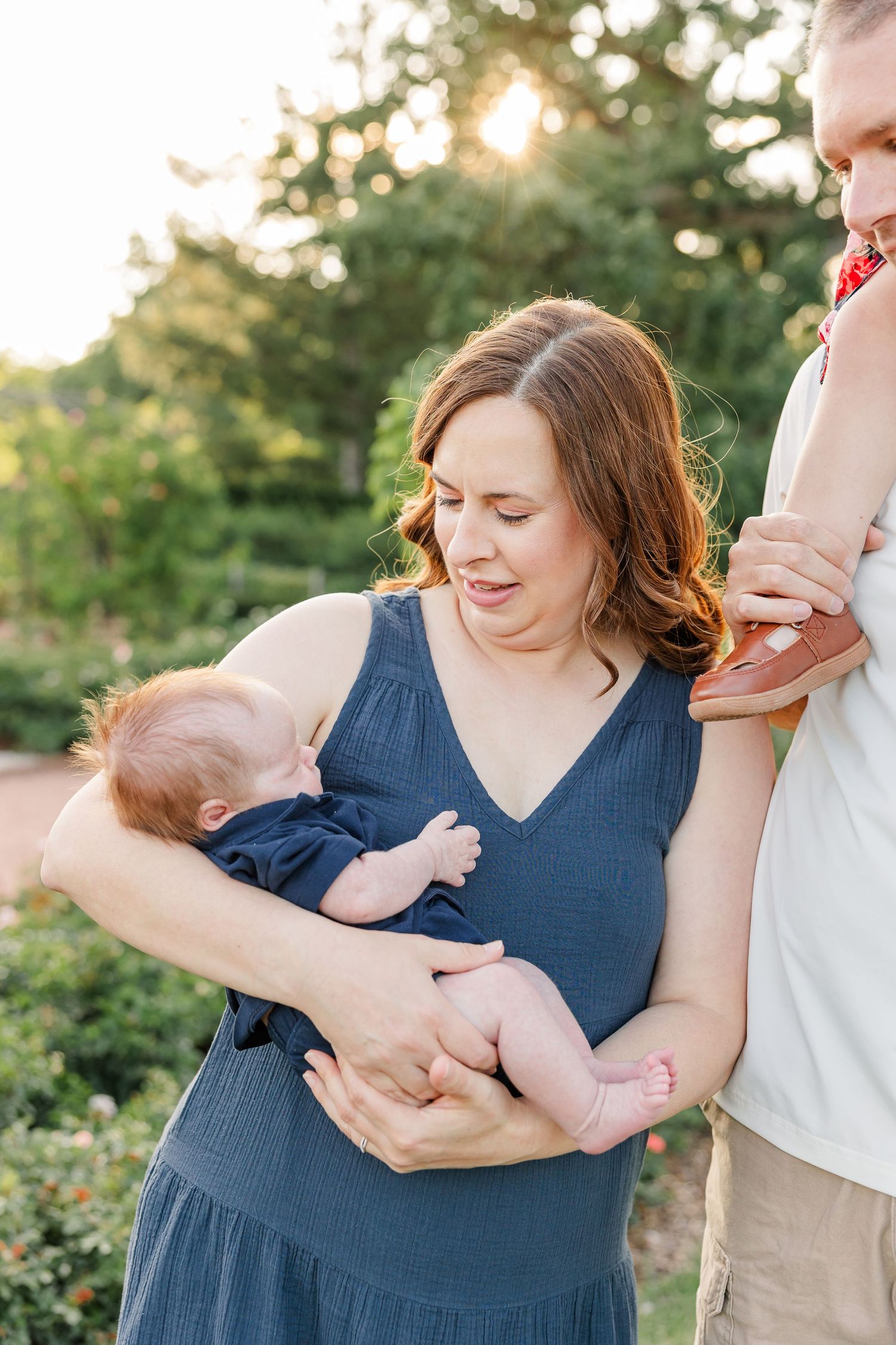 Hilary smiling down at newborn Lincoln while holding him outdoors during their summer family session.