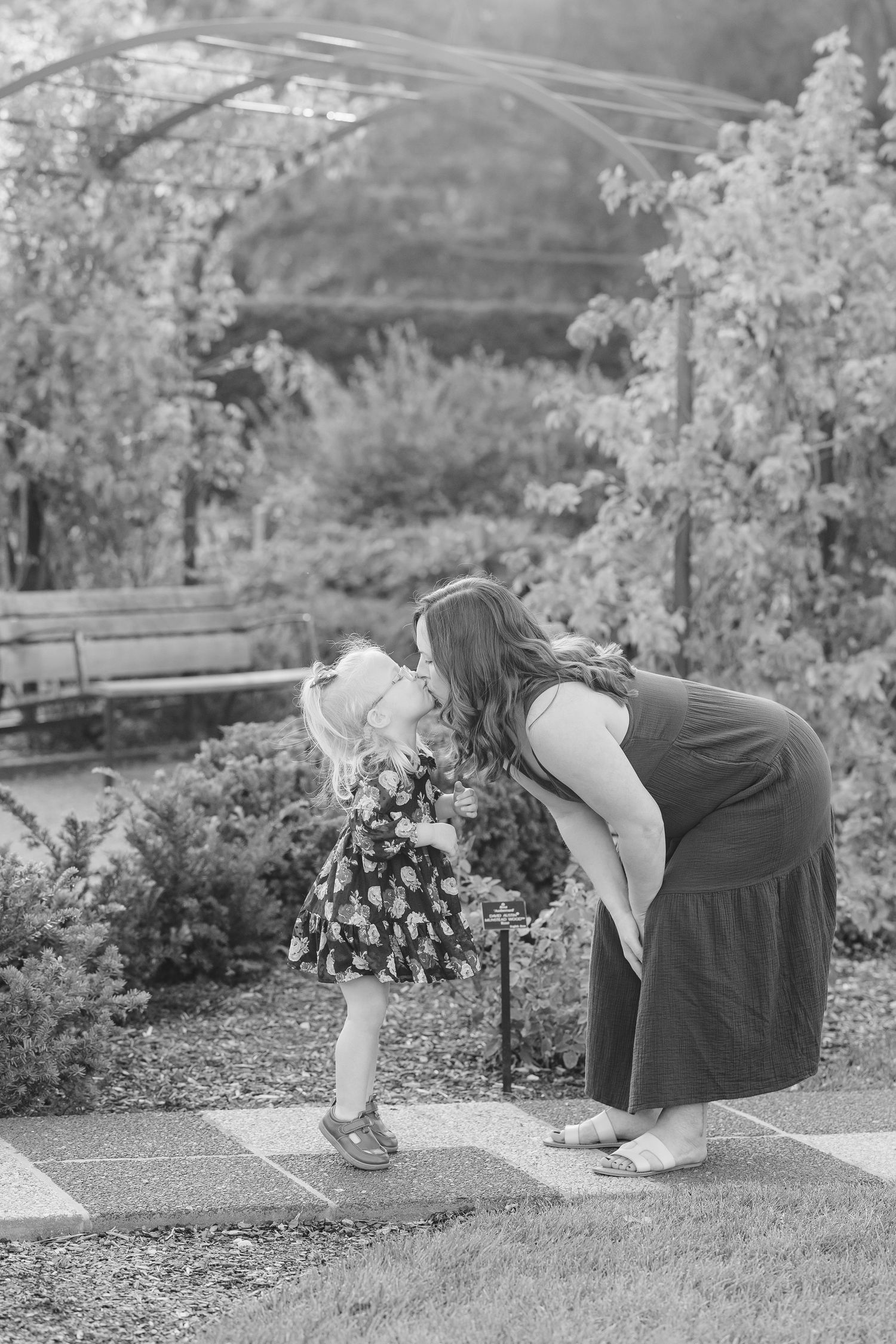 Black-and-white portrait of Hilary kissing Caroline beneath a flower-covered arch at Cantigny Park.