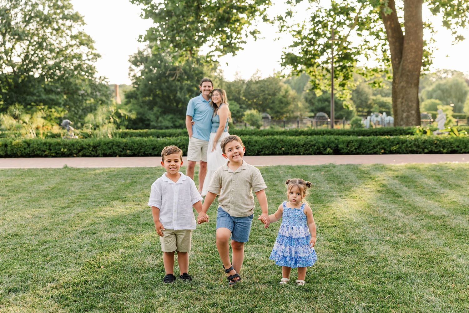 Happy family with parents and three young children holding hands and walking together on green lawn in garden.