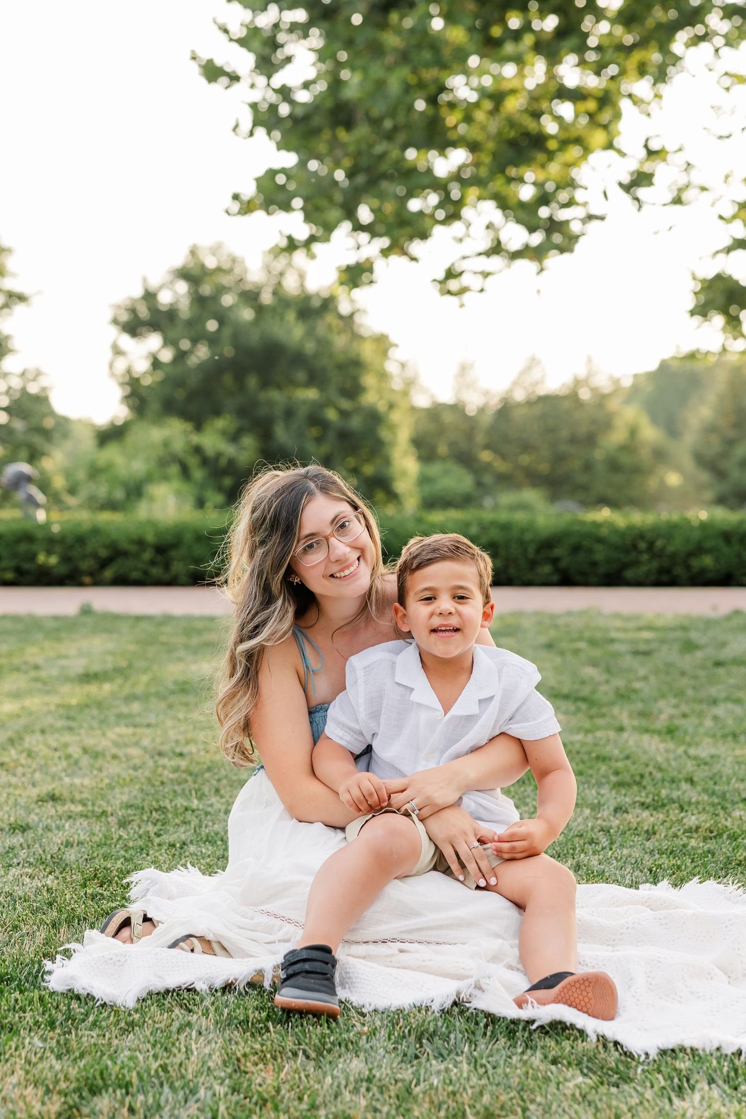 Smiling mother in white dress sitting on blanket in garden with young son in white shirt on sunny day.