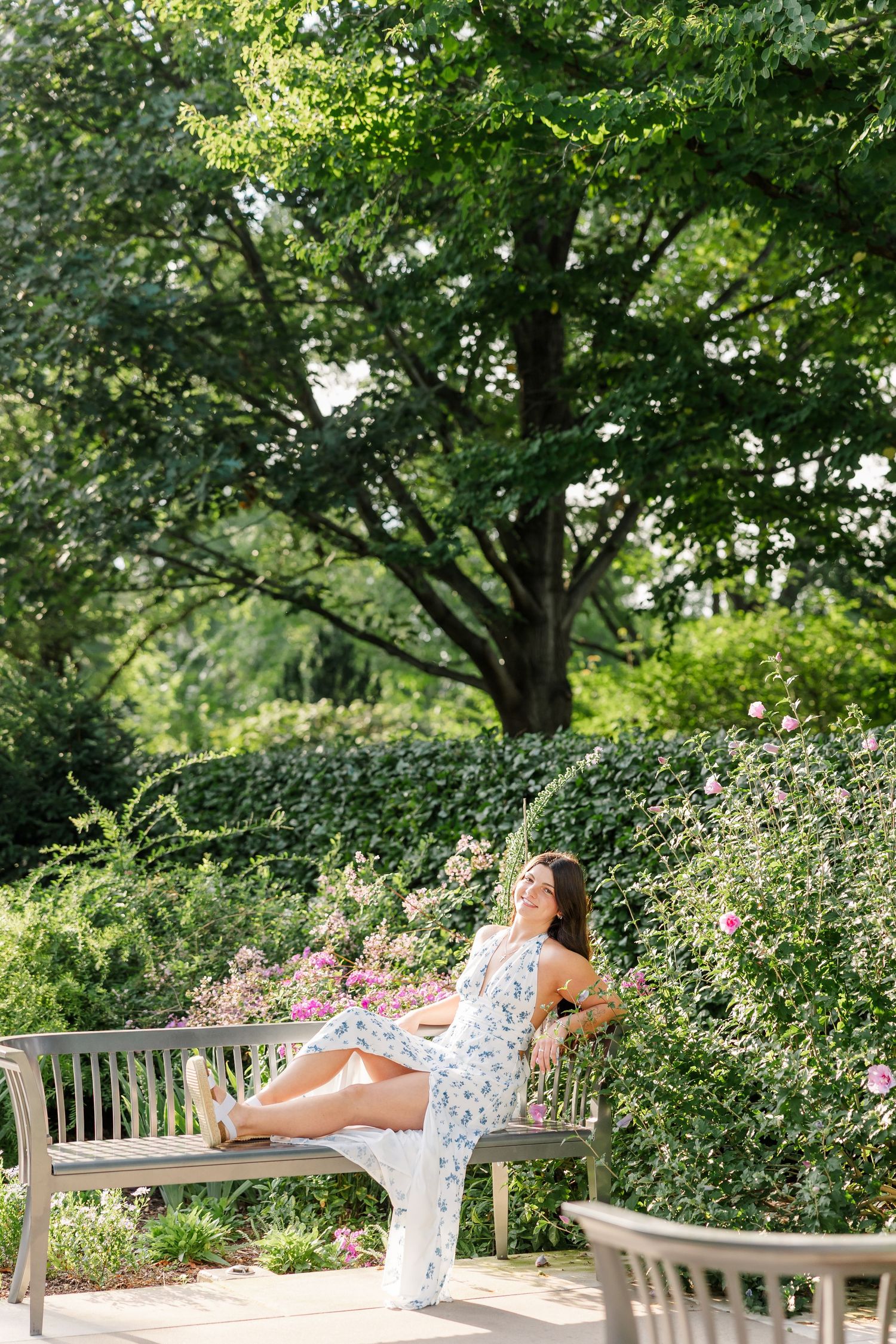 Elegant woman in floral maxi dress relaxes on garden bench surrounded by blooming roses and greenery.