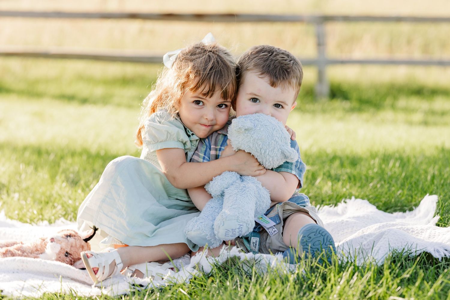 Two young children hugging on a blanket in green grass, sharing a sweet moment with a stuffed animal toy.