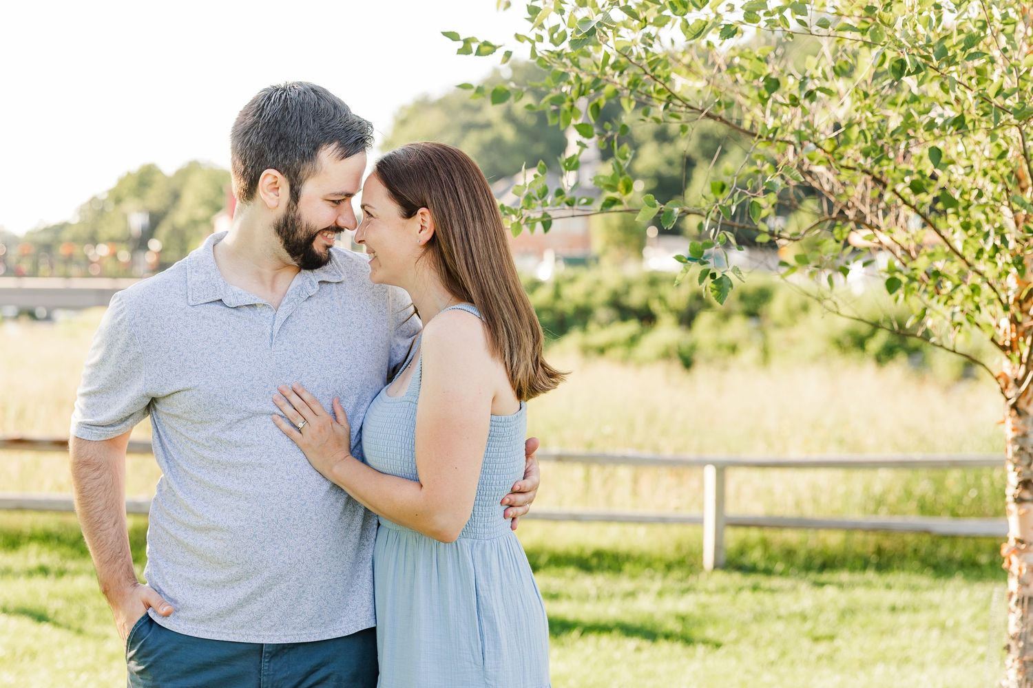 A couple shares a romantic moment outdoors, smiling at each other near a tree with a sunny field behind them.