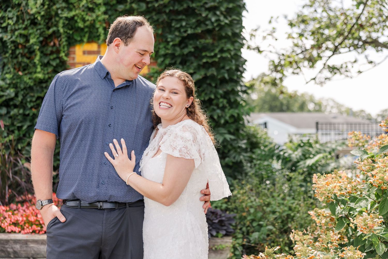Joyful couple posing outdoors in a garden, bride showing off engagement ring while laughing.