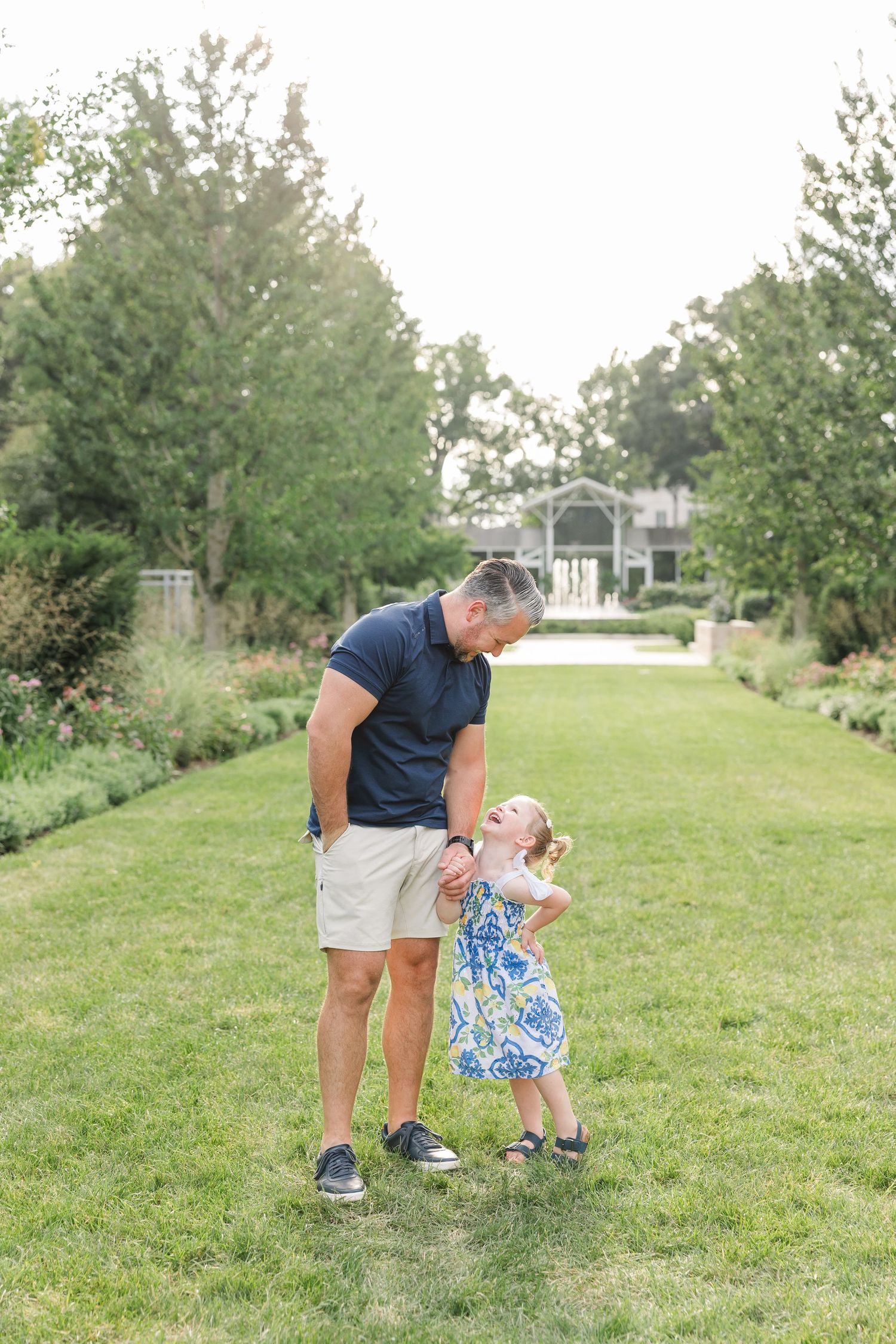 Father in navy polo bends down to interact with his young daughter in a blue floral dress on a green lawn.