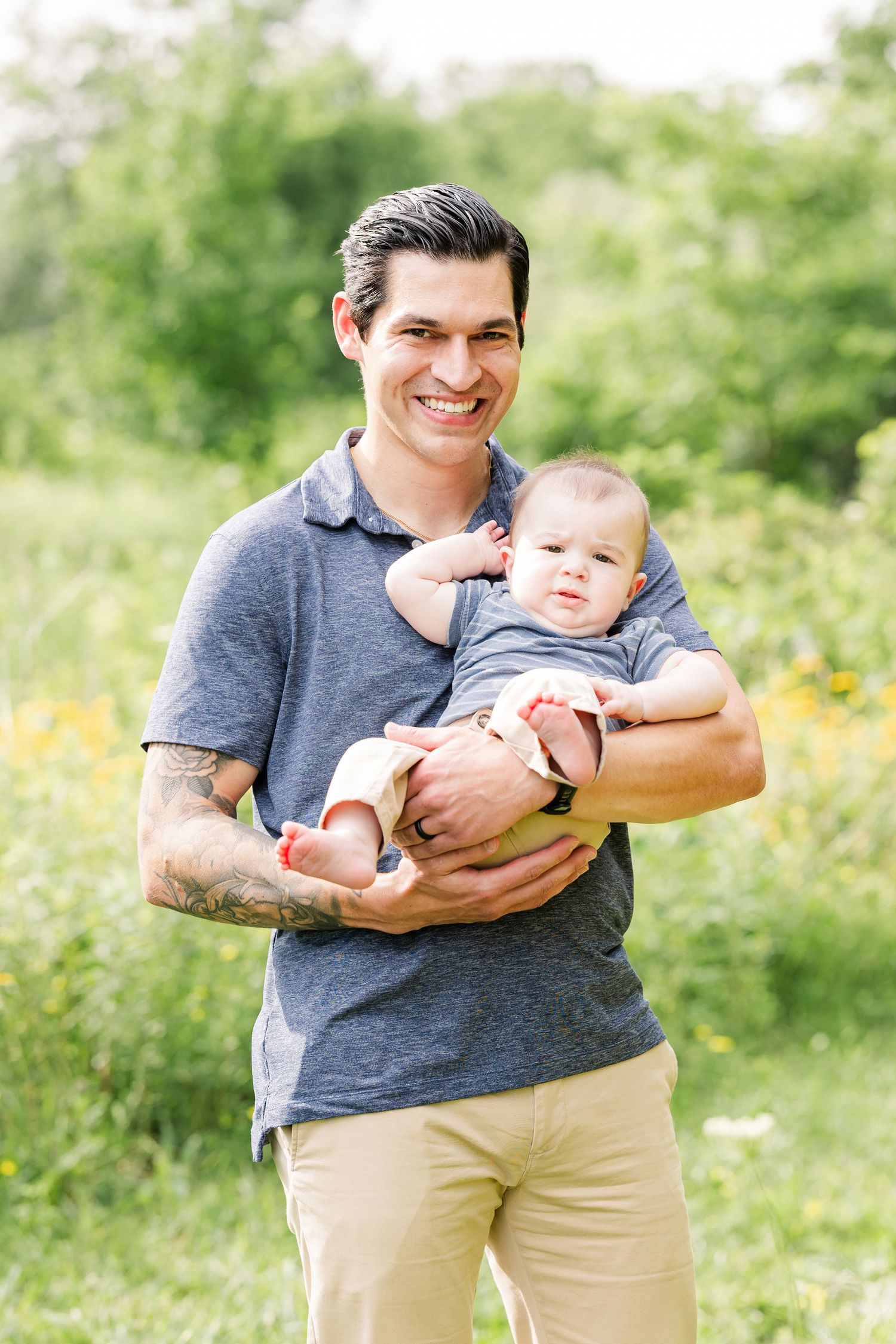 A smiling man with tattoos holds his baby in a sunny green outdoor setting while wearing a gray polo shirt.
