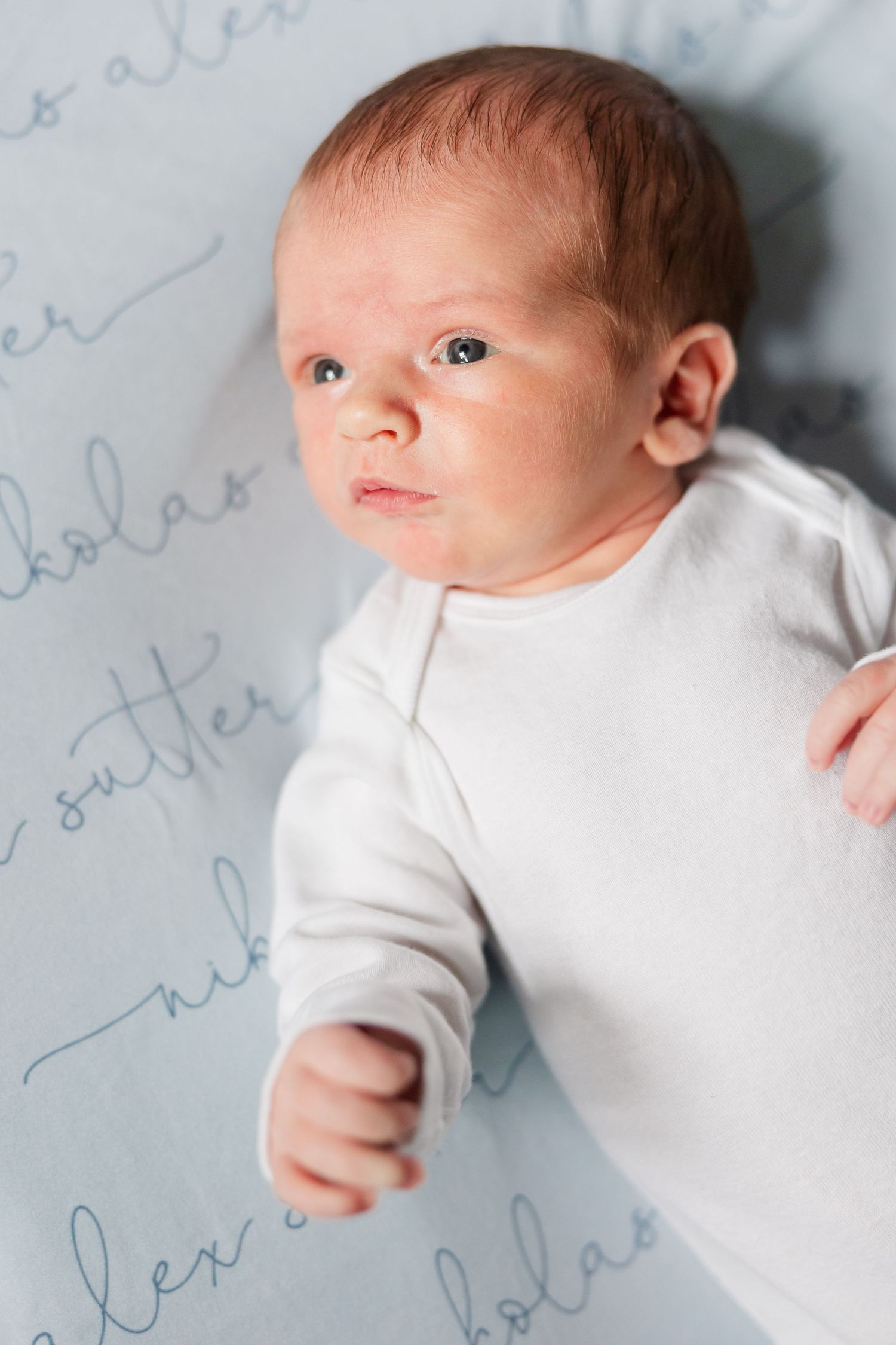 Alert newborn baby in white onesie lying on personalized blue blanket with name script print.