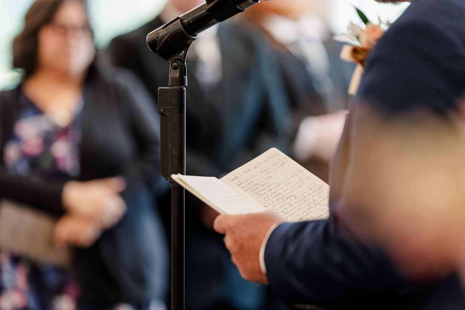 Person in a navy suit reads from a folded paper at a microphone while guests stand blurred in the background at an outdoor ceremony.