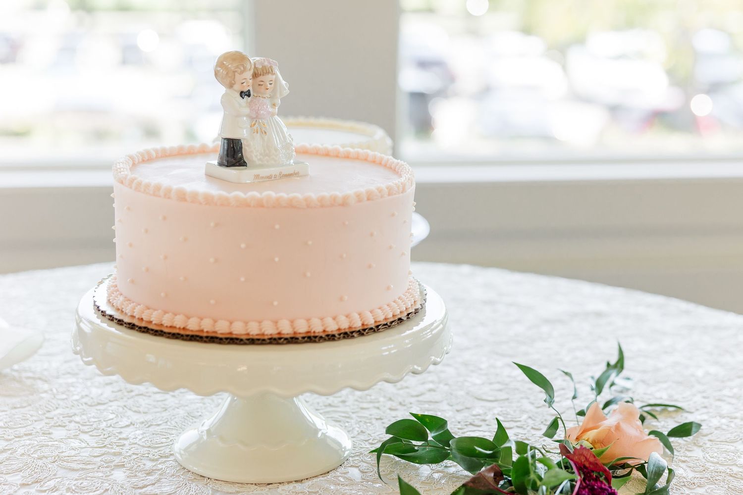 A pink buttercream wedding cake topped with a bride and groom figurine sits on a white cake stand beside fresh floral greenery.