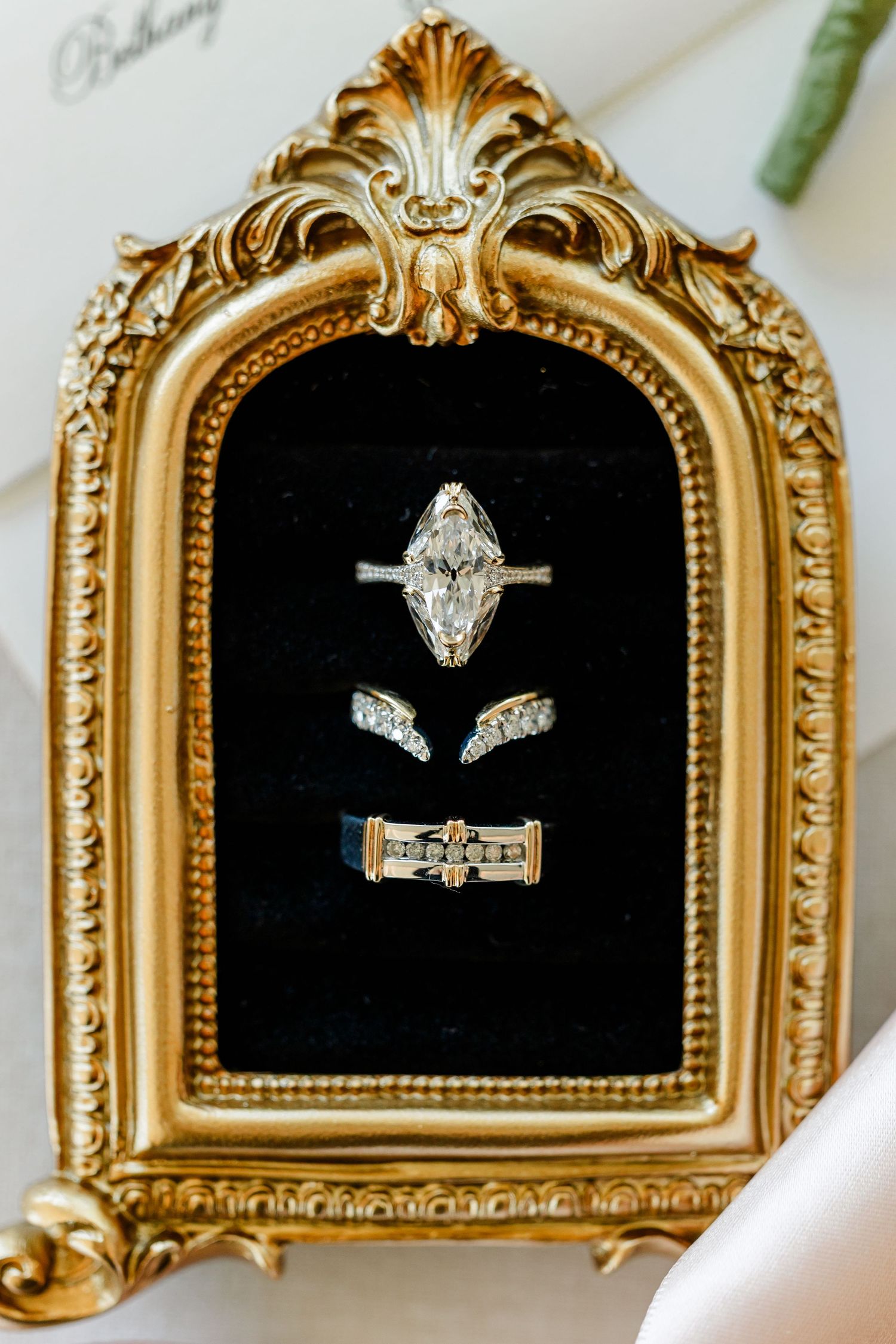 Marquise diamond engagement ring and two wedding bands displayed on black velvet inside an ornate gold baroque frame.