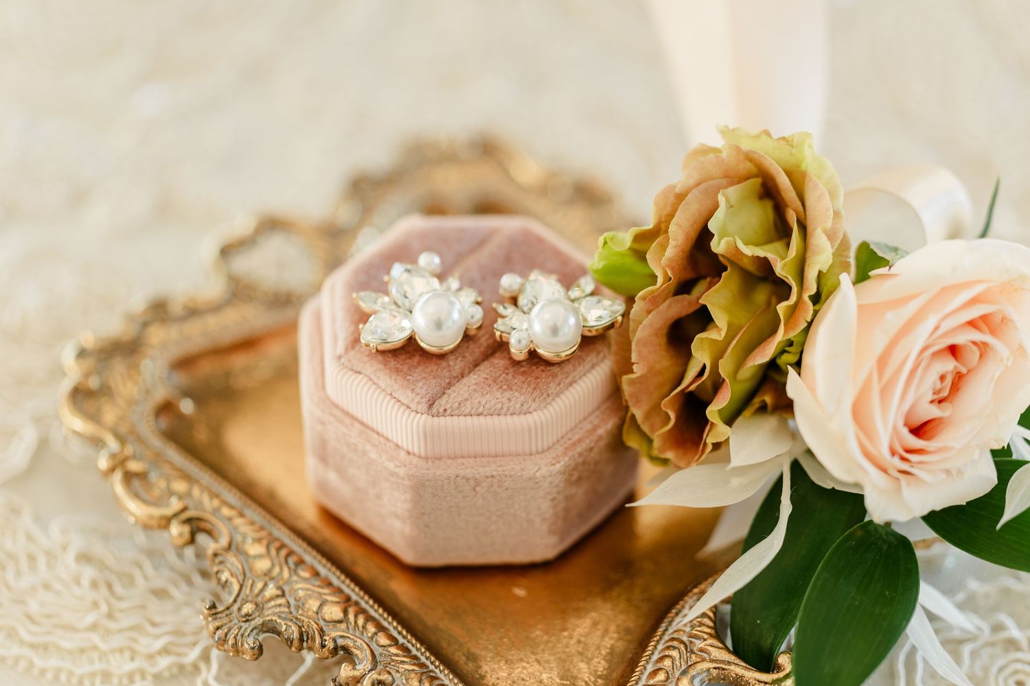 Pearl and crystal floral bridal earrings on an octagonal blush velvet ring box atop an ornate gold tray beside a peach rose bouquet.