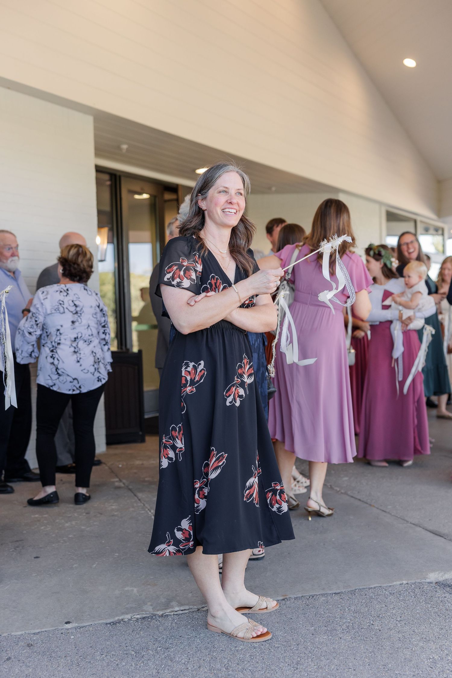 Smiling woman in a black floral dress holds a white ribbon wand outside a building while wedding guests gather behind her.