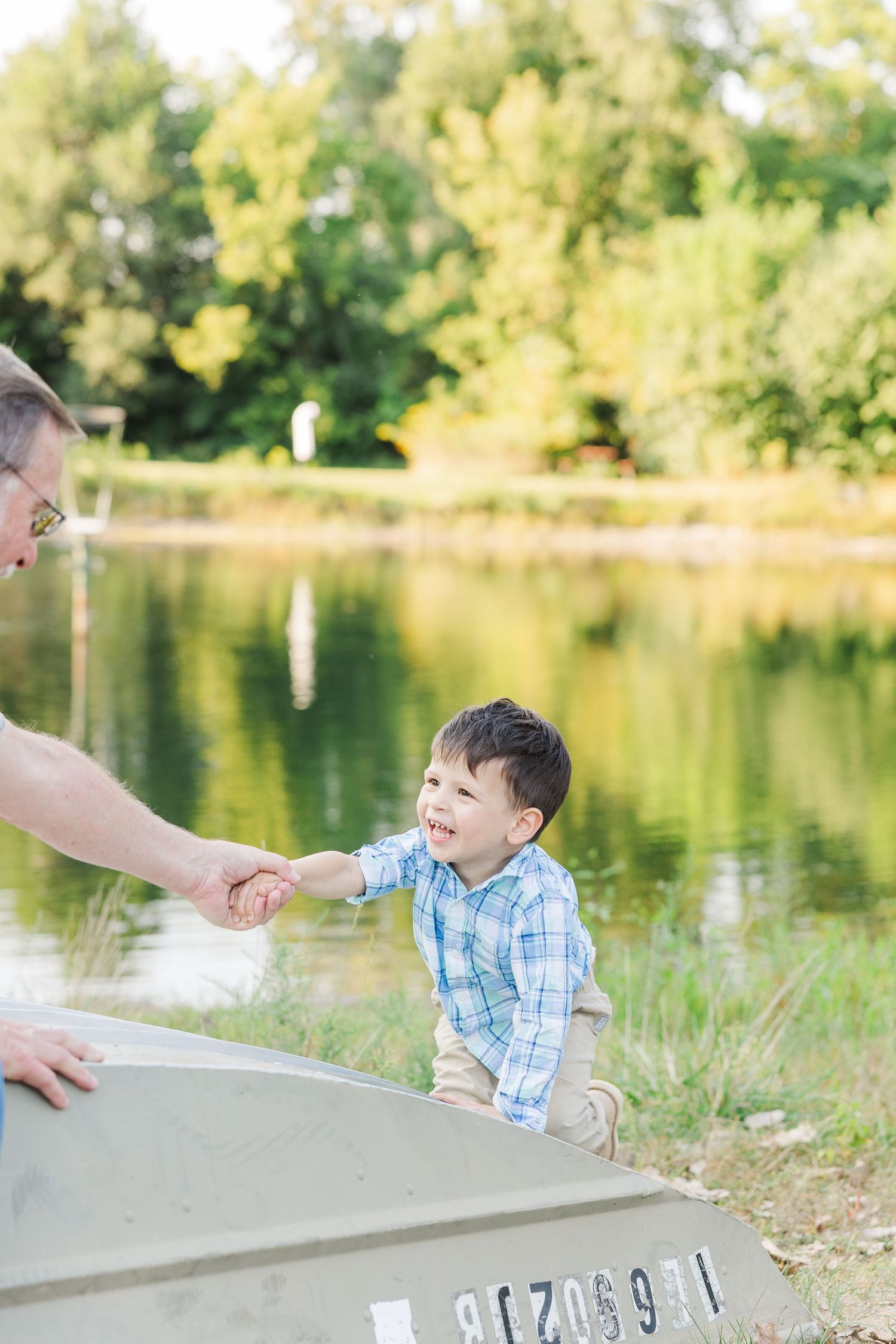 Child plays outdoors near calm lake on sunny summer day in blue plaid shirt while adult hand reaches in frame.Sorg family portraits at Shamr