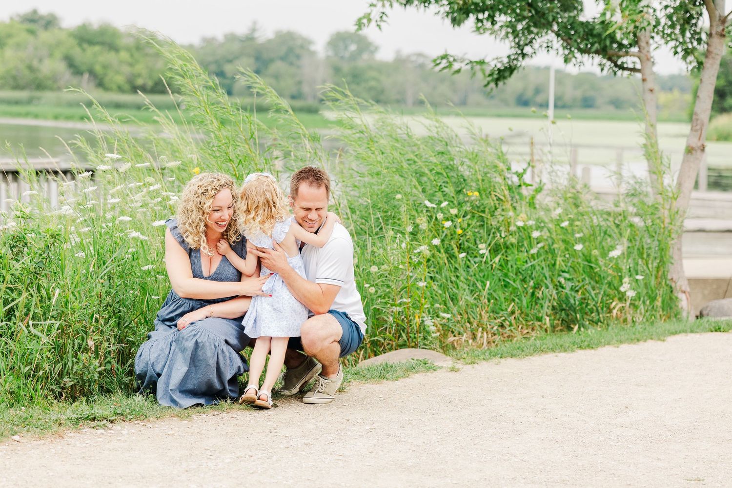 A touching family moment captured on a dirt path by a serene lake surrounded by green foliage.