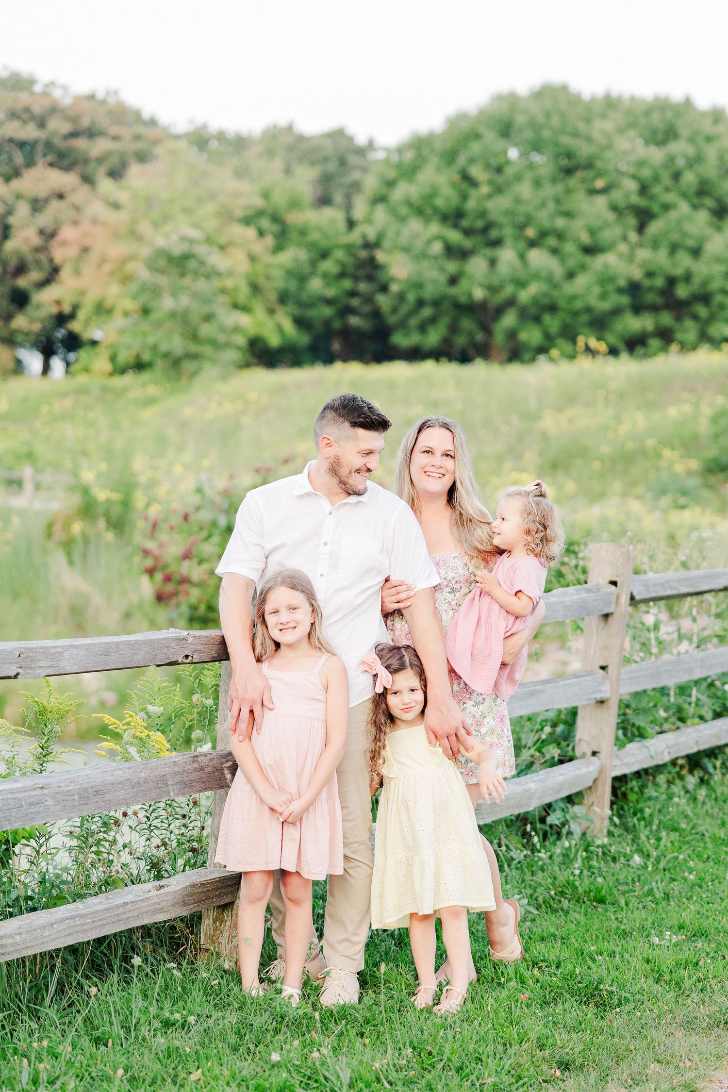 Family poses together by rustic wooden fence in summer meadow wearing coordinated pastel outfits.