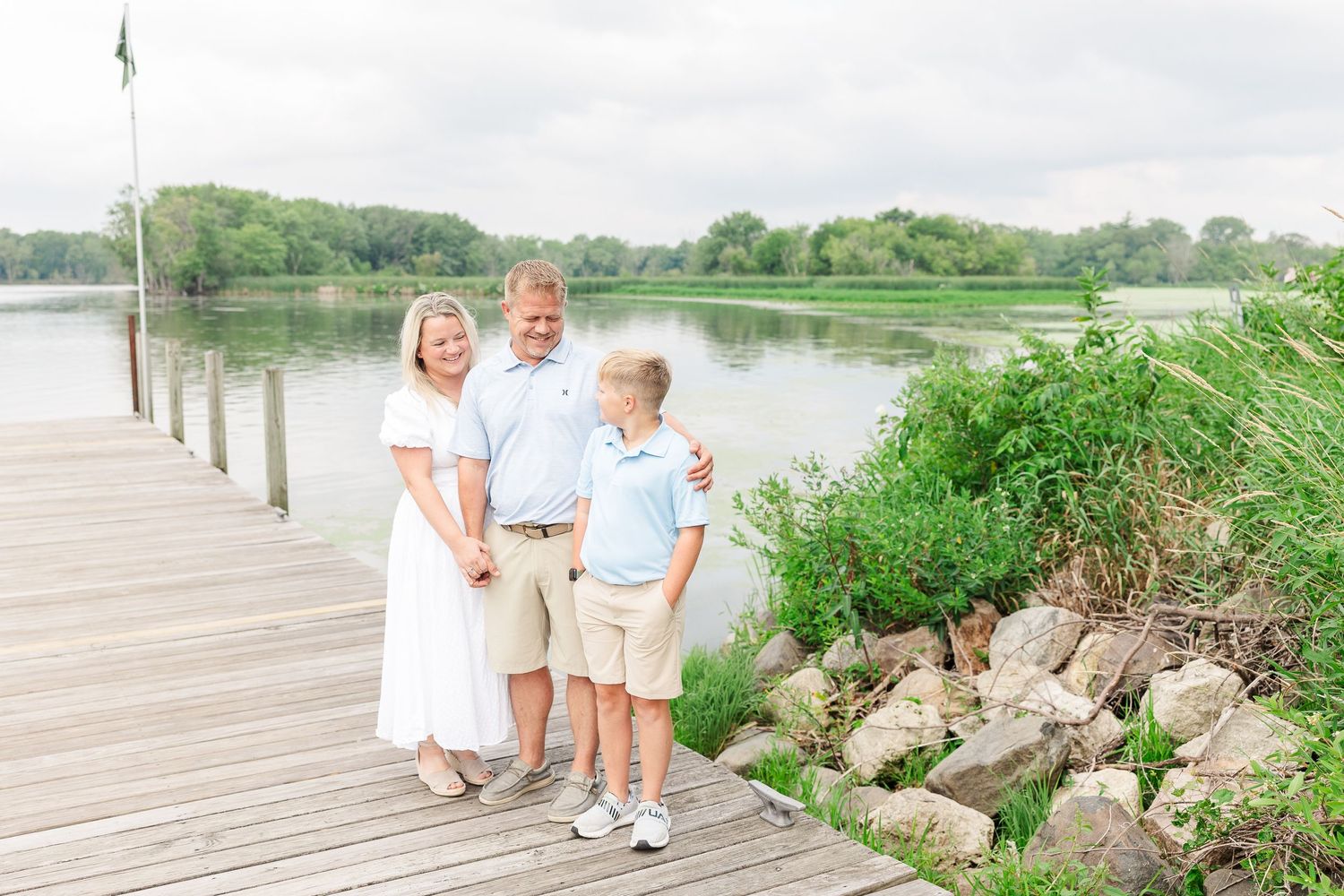 Family standing together on a wooden dock by a serene lake surrounded by green trees on a cloudy day.