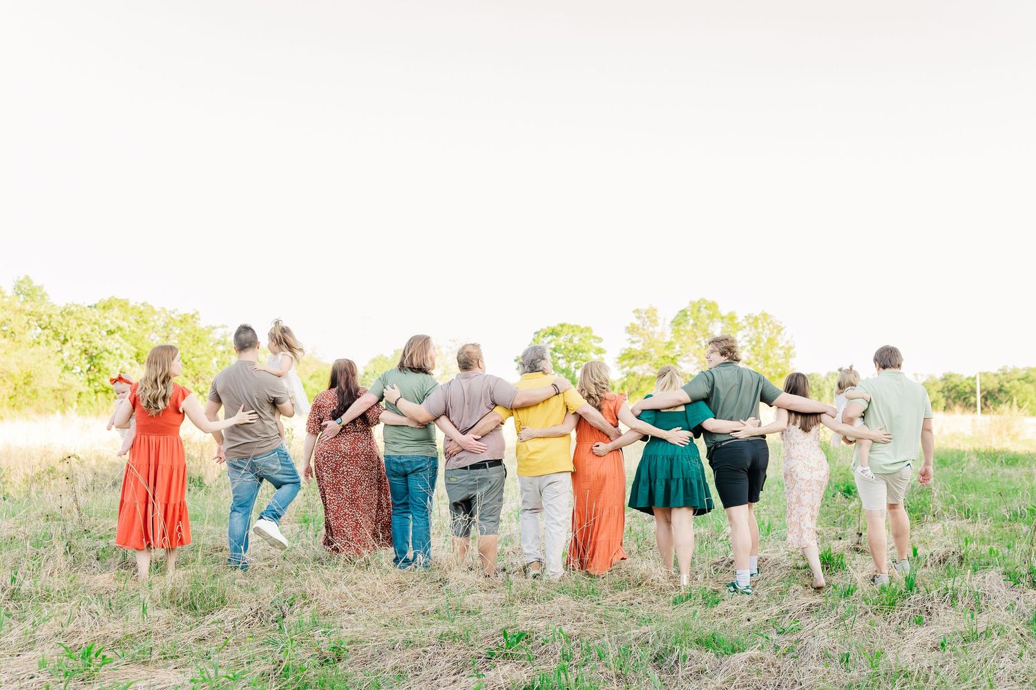 Friends lined up with arms around each other walking away from camera on grassy field at sunset.