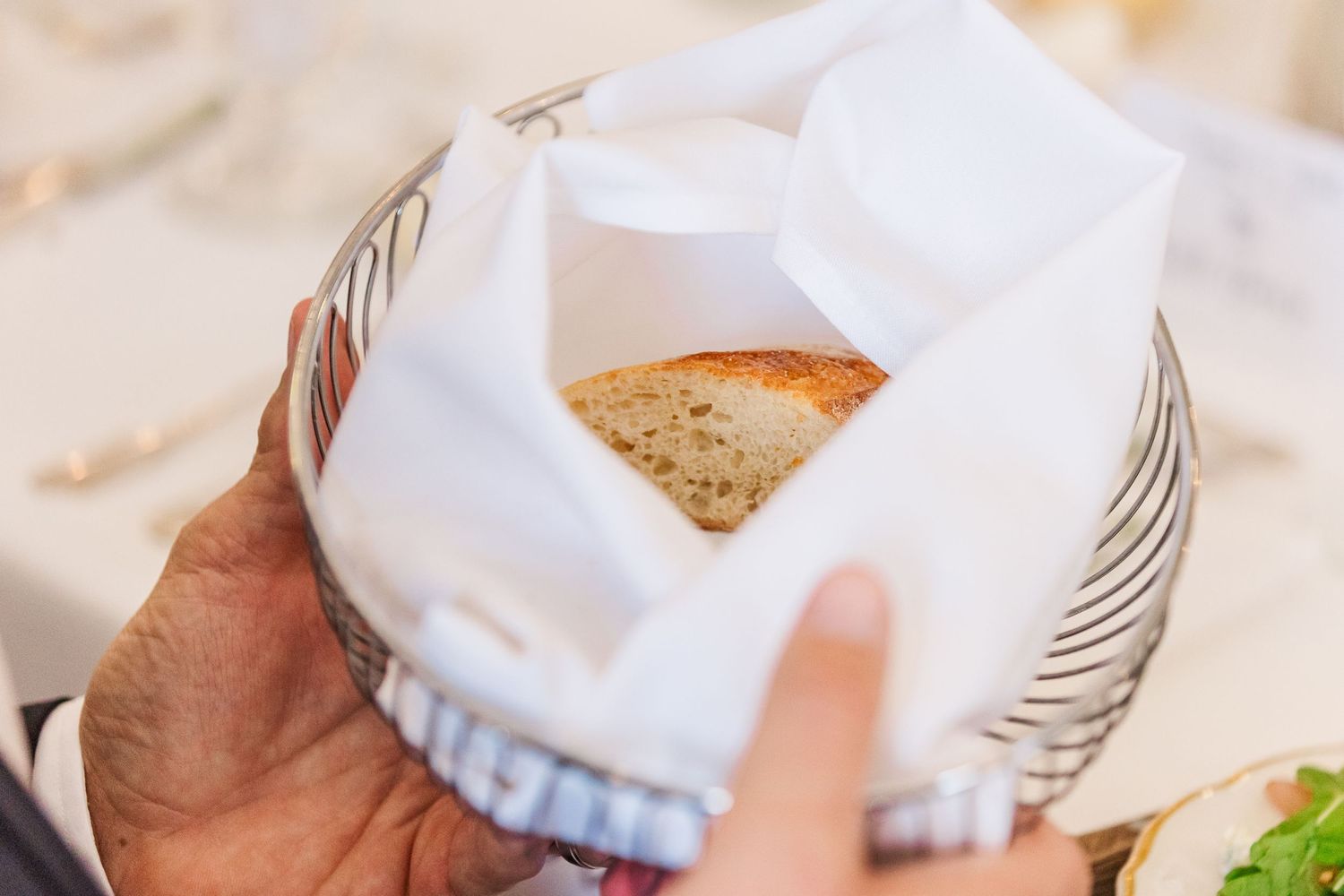 Hand holding a piece of bread wrapped in white cloth or napkin at what appears to be a formal dining setting.