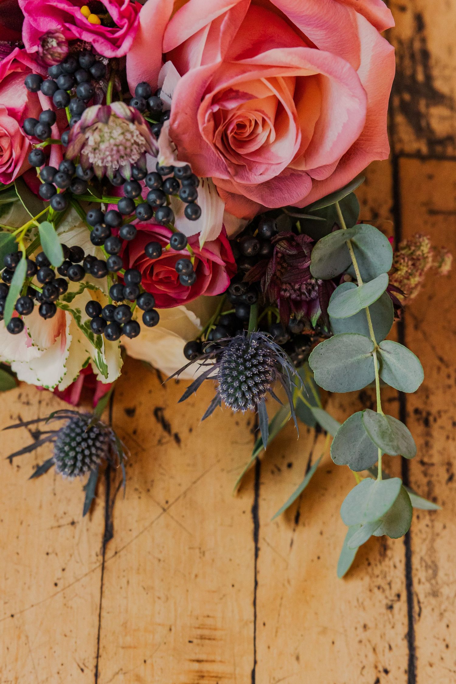 Pink and red roses with blue thistle and eucalyptus arranged on a rustic wooden surface.