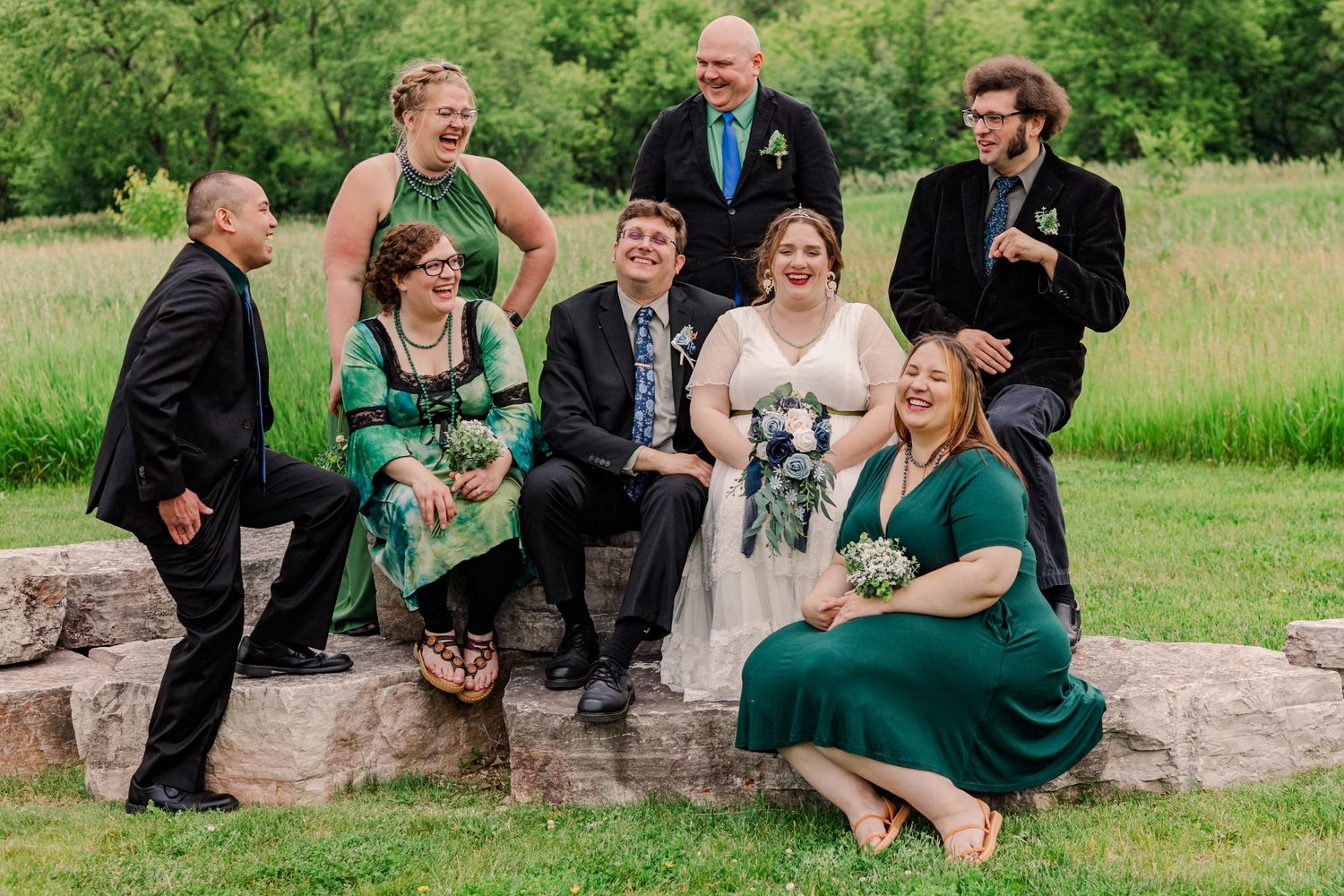 Wedding party members sit casually on rocks while posing for photos outdoors.