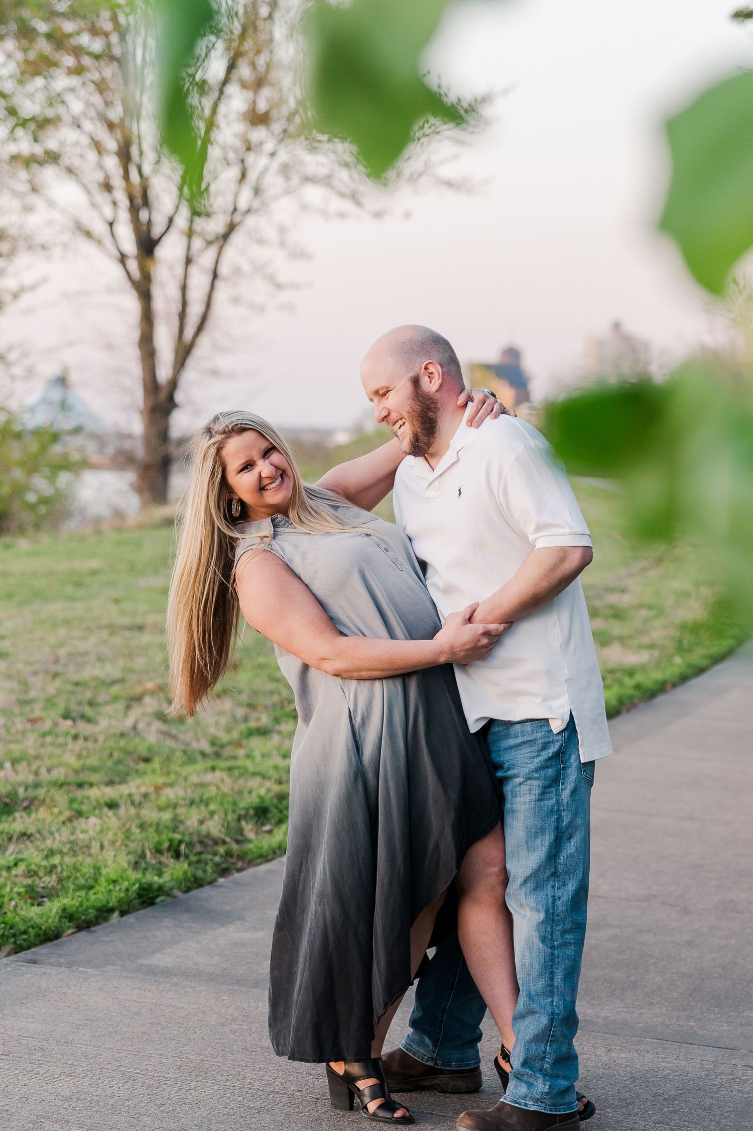 A romantic couple embraces on a paved path surrounded by green trees and grass in evening light.