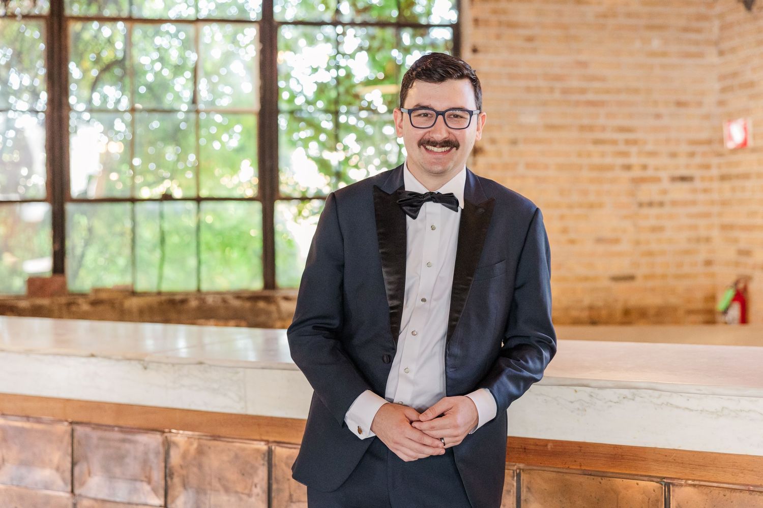 A person in formal attire smiles warmly in front of a rustic brick wall and bright windows.
