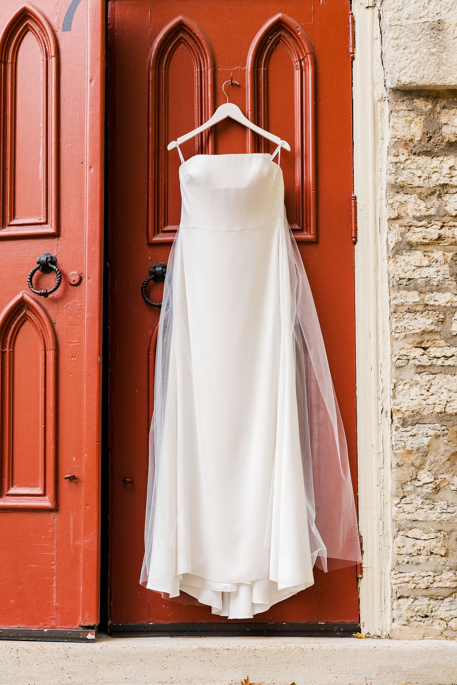 White wedding dress hanging on a wooden hanger against a red church door with Gothic panels.