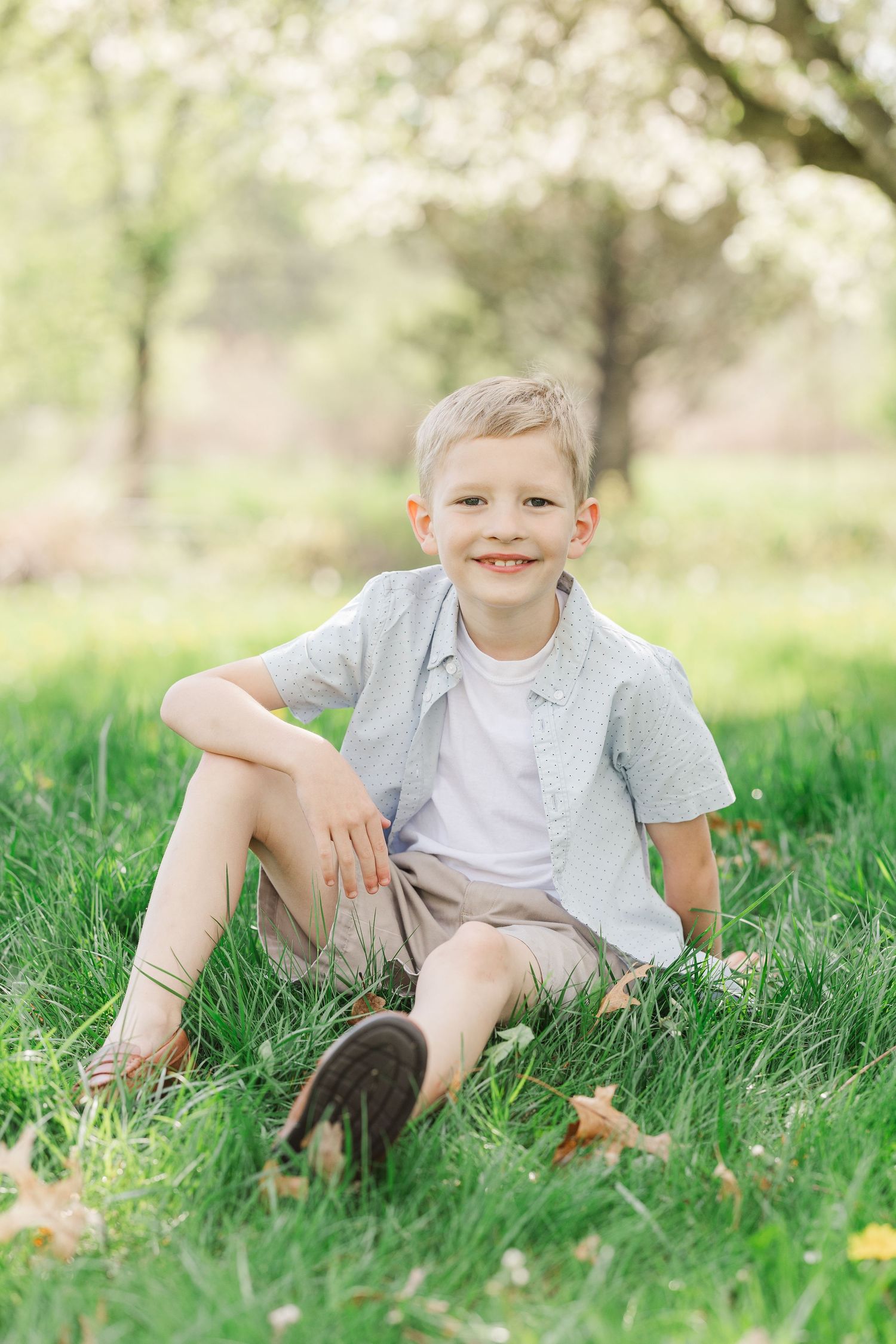 A young child sits casually in green grass under blooming spring trees.