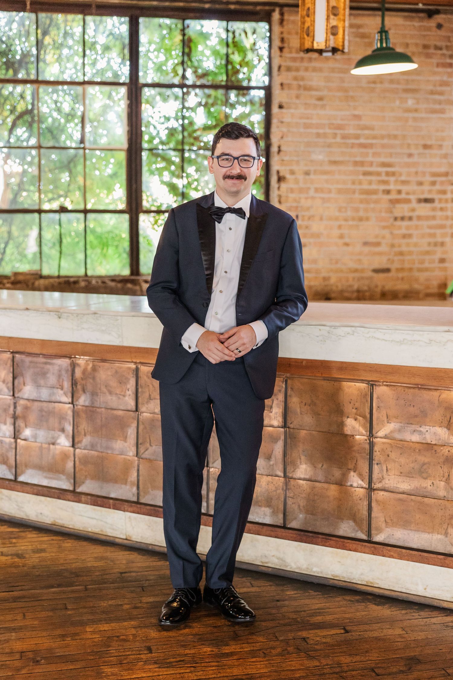 A formal portrait of a person in a tuxedo standing against a rustic brick wall and wooden bar.