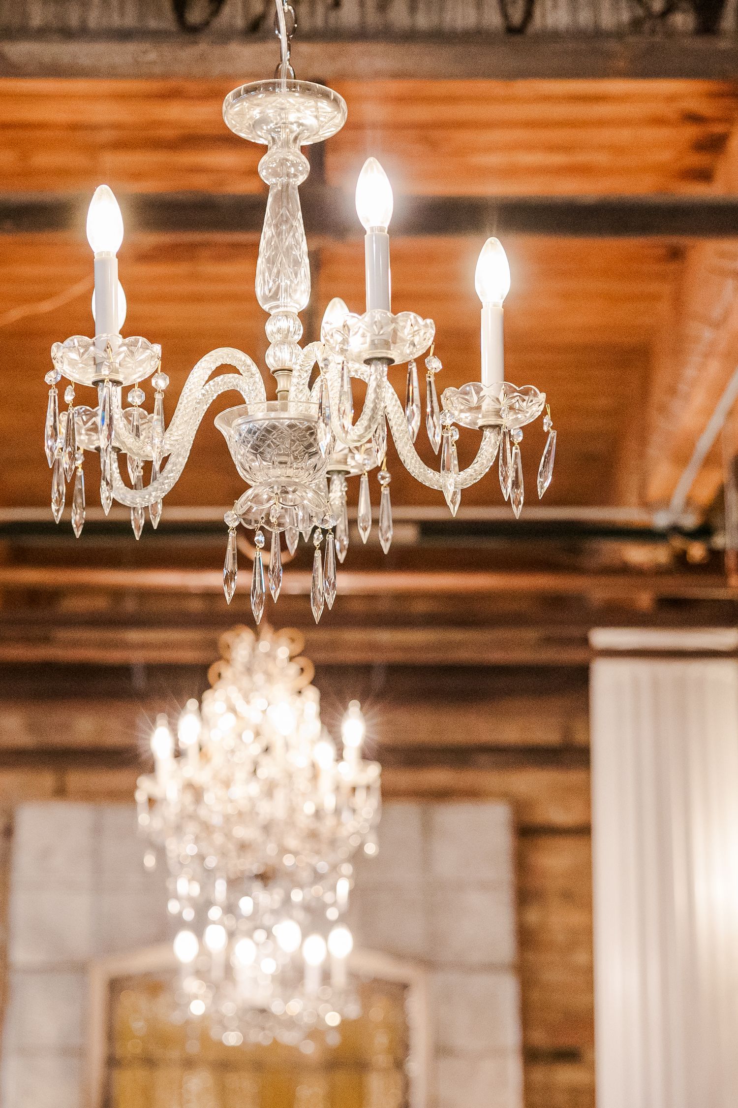 A delicate white crystal chandelier with multiple arms illuminated against a wooden ceiling.