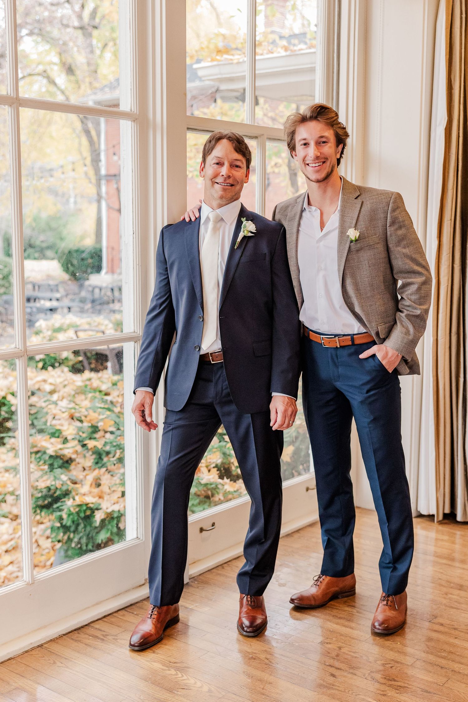 Two men in suits pose casually by large windows with autumn foliage visible outside.