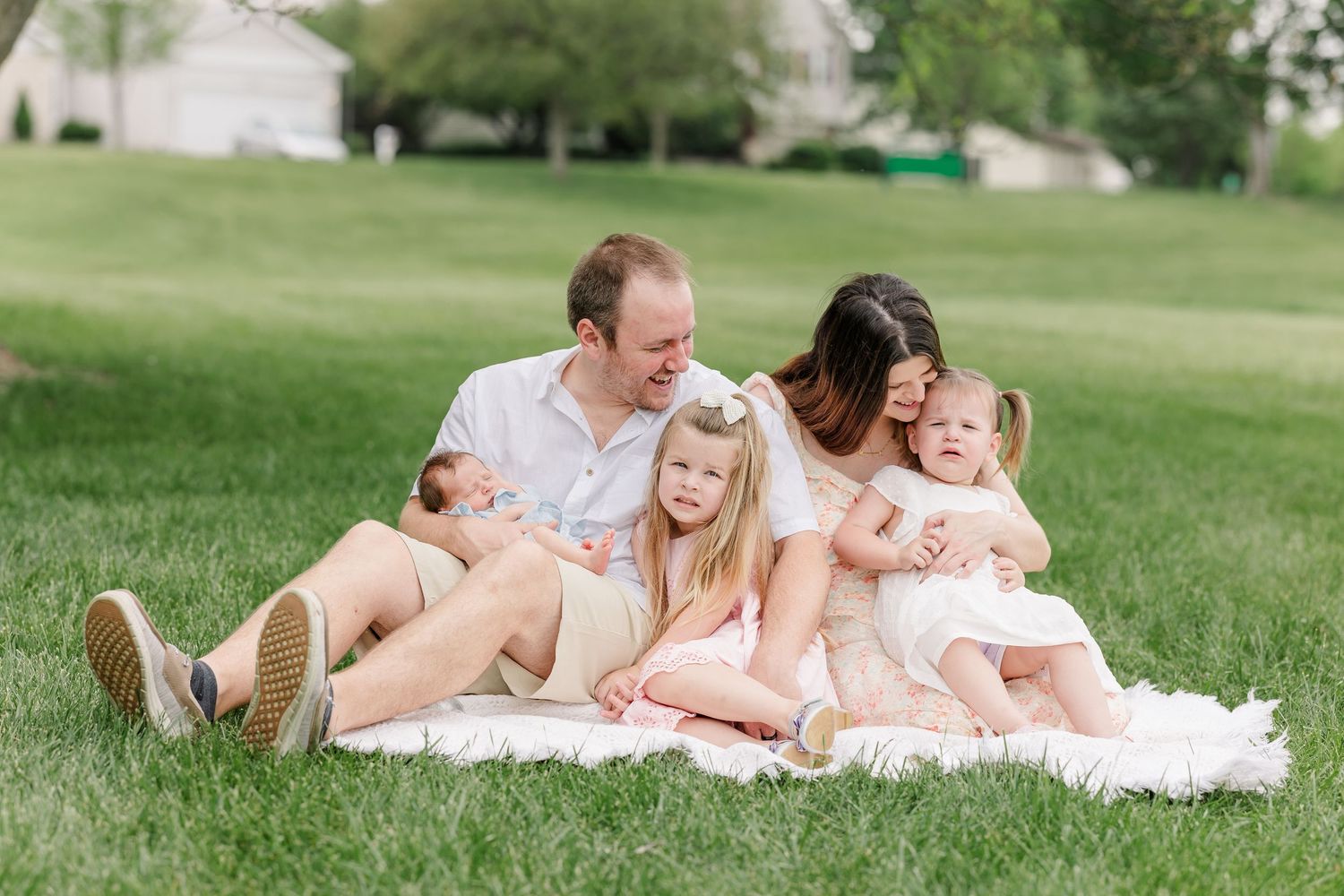 Family of five sitting on a white blanket in a lush green park, parents cuddling three young children on a sunny day.