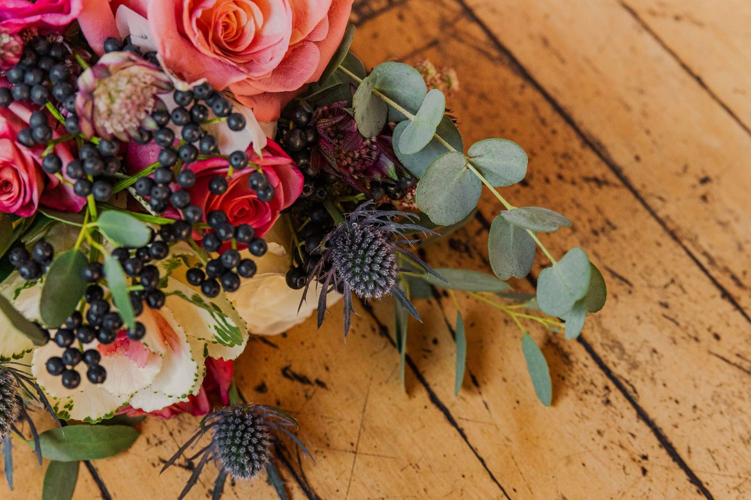 Vibrant wedding bouquet with pink roses, blue thistle, and eucalyptus on wooden surface.