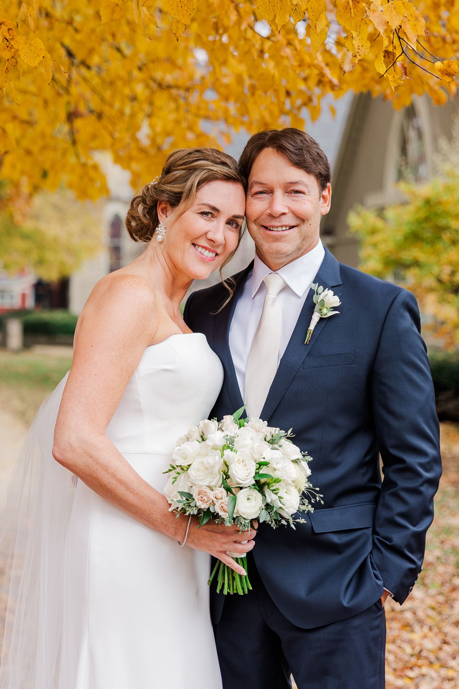 Wedding couple smiling at camera with vibrant yellow autumn leaves overhead.