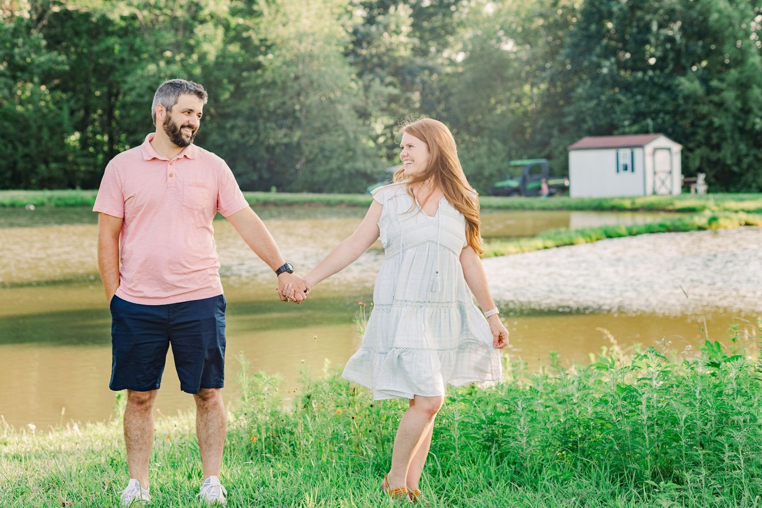 Couple holding hands while standing in grassy field near a lake at sunset.