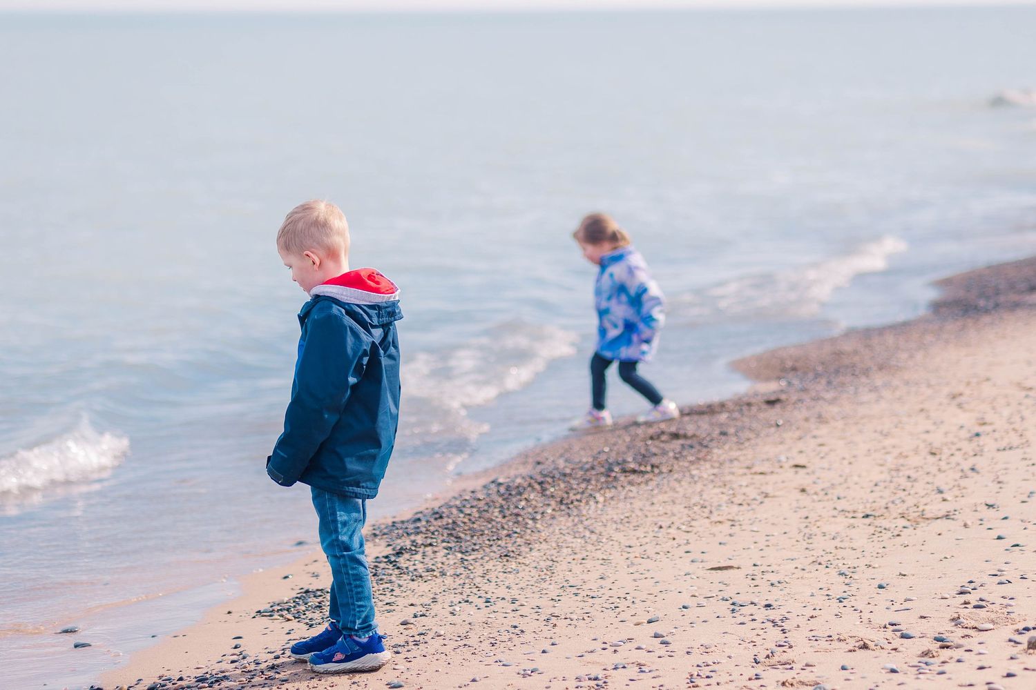Two children in winter coats explore rocky beach along water's edge.